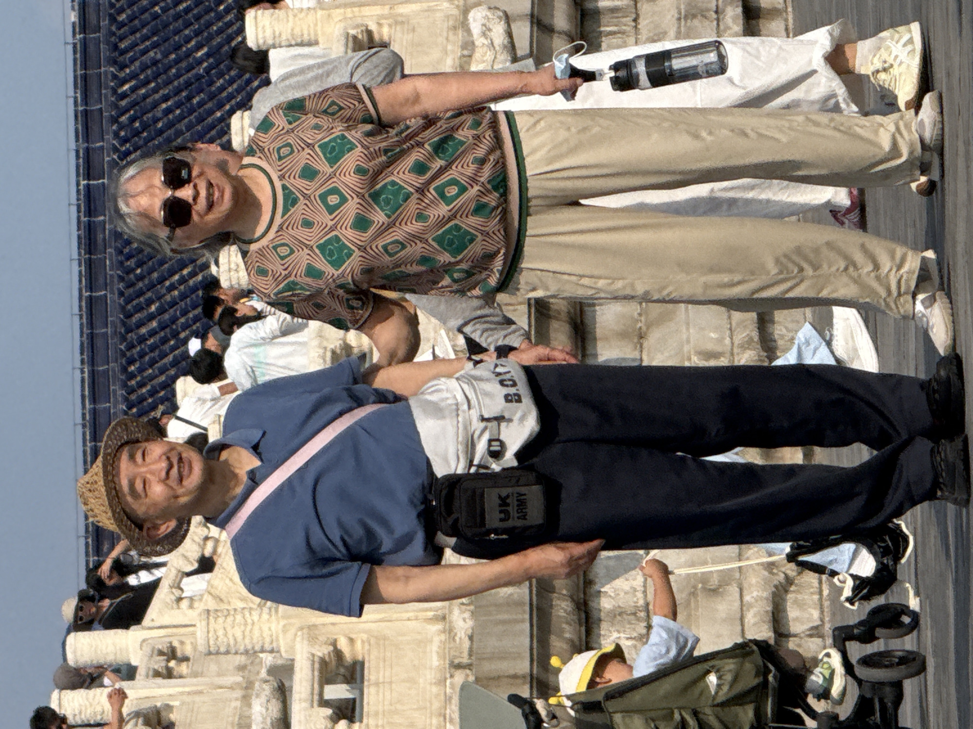 An older Asian man wearing a straw hat, blue shirt, and dark pants stands in the foreground, slightly to the left of center. He has a pink strap across his chest and a black pouch with "UK ARMY" printed on it hanging from his belt. To his right, an older Asian woman wearing sunglasses and a patterned green and tan shirt with beige pants stands. Her right hand holds a black water bottle with a white label. Behind them, a child in a yellow hat and a green backpack is in a stroller. The background shows stone structures and a dark blue roof, suggesting a historical site. The scene appears to be outdoors in a public area, possibly a tourist attraction in Dongcheng District, China, given the architectural style.