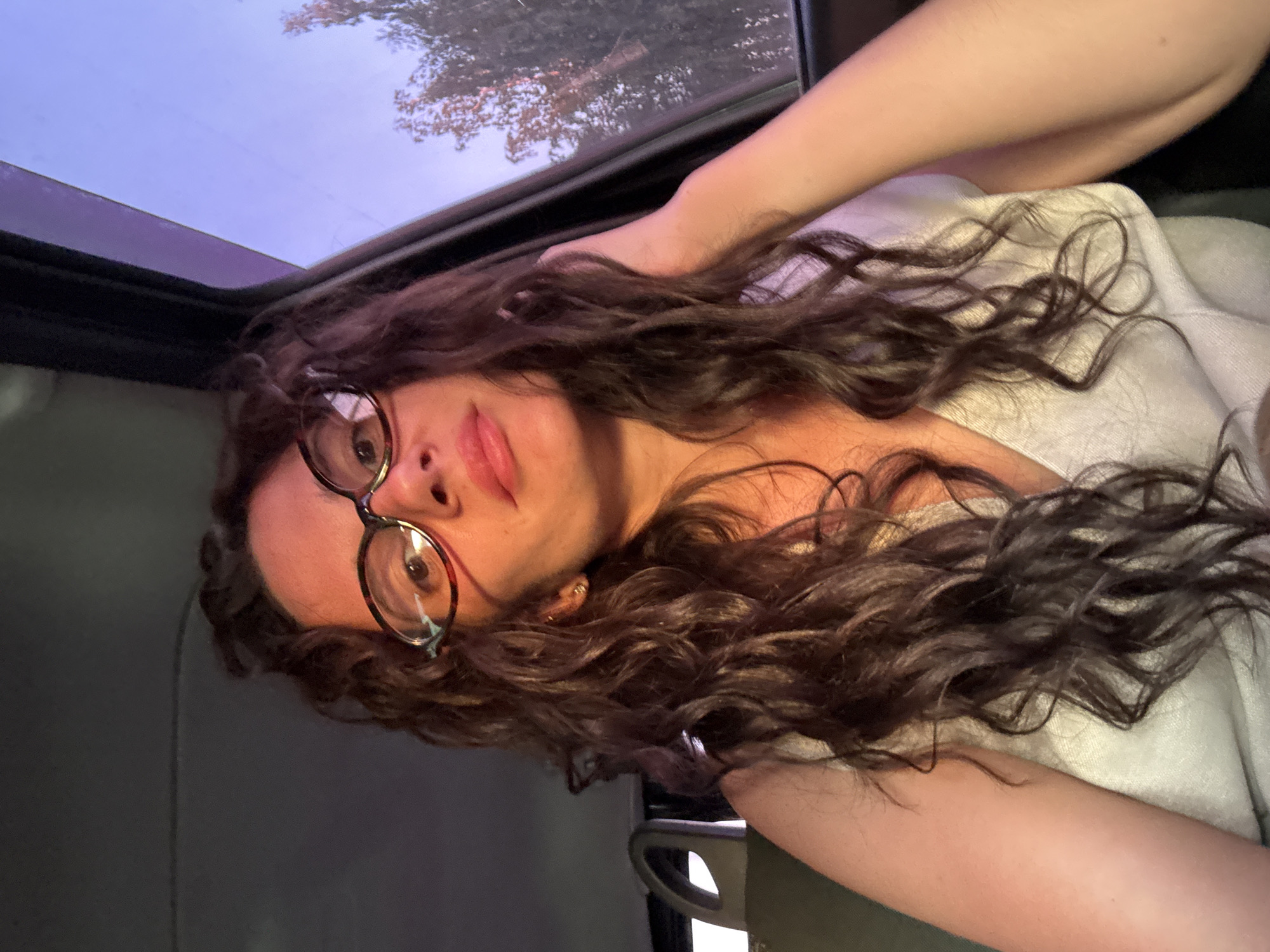 The image shows a woman with curly brown hair and glasses lying down, possibly in a car with a sunroof, judging by the visible frame and sky. She is wearing a light-colored garment. The environment visible through the sunroof appears to be a twilight sky with tree branches. There is no information in the image or provided context to indicate that this image was taken in the city of Chaikovsky, Russia.