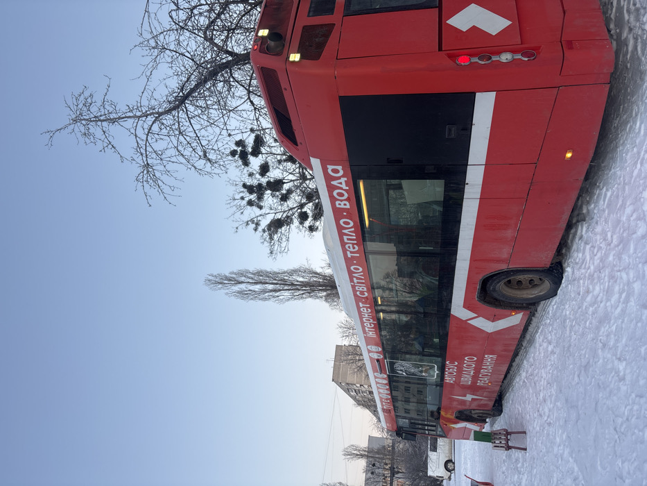 This image depicts a snow-covered urban environment in a city in Ukraine, under a clear, pale blue sky during daylight. The primary subject is a stationary, red and white articulated city bus, positioned with its rear angled towards the viewer's right. The bus displays Ukrainian text: "ІНТЕРНЕТ • СВІТЛО • ТЕПЛО • ВОДА" (Internet • Light • Heat • Water) along its upper white stripe, and "АВТОБУС ШВИДКОГО РЕАГУВАННЯ" (Rapid Response Bus) accompanied by a lightning bolt symbol below the windows. A dark crest or emblem is also visible on the side. The bus's lower body and wheels show accumulation of dirt and snow. Indistinct figures are discernible through the tinted windows, suggesting occupants. One red brake light on the rear of the bus is illuminated. To the left of the bus, a small, wooden bench, partially covered in snow, is present. In the background, bare deciduous trees, some with visible clumps of parasitic growth (likely mistletoe), rise against the sky, and multi-story grey residential buildings, characteristic of post-Soviet architecture, are visible. A white utility van is partially visible parked further to the left in the snowy landscape.