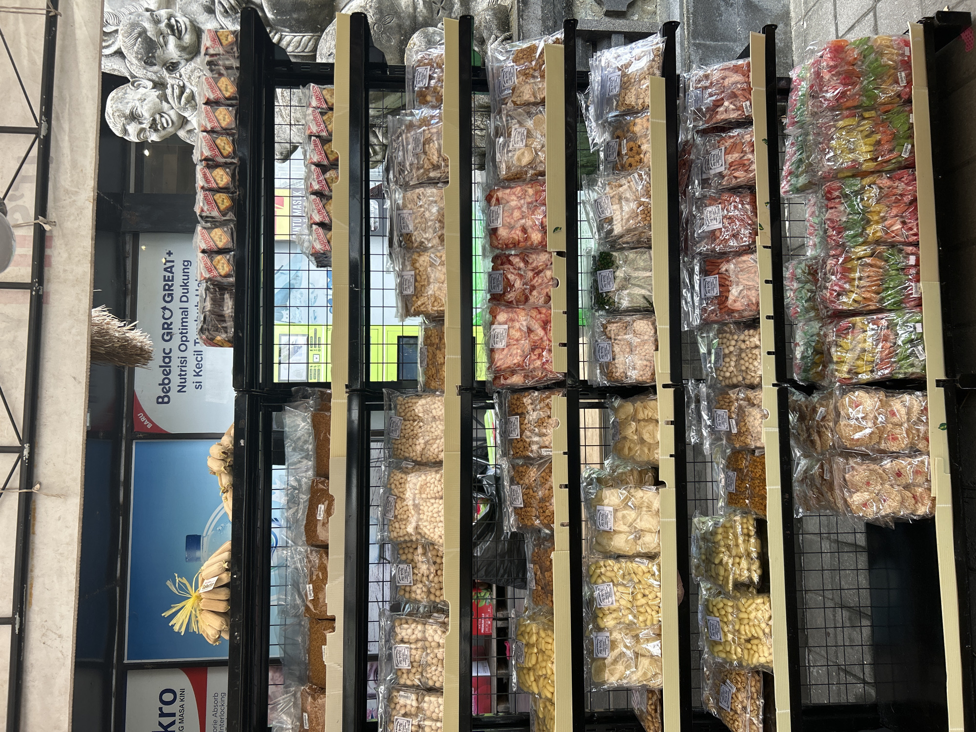 This image captures a detailed view of a retail display in Sukawati, Indonesia, likely at a market or souvenir shop. The focus is on shelves stocked with a variety of packaged snacks and goods.
The display shelves are organized with black metal framing and light-colored, possibly wooden, shelves. The top shelf, partially visible, features promotional posters and what appears to be bundles of dried goods. Below this, several shelves are filled with numerous transparent plastic packages of various food items. These include nuts, puffed snacks, and what seem to be dried fruit or candy pieces in vibrant colors. Many packages have labels with text, though it is not clearly legible.
In the background, a large stone sculpture of two smiling children is visible, adding a cultural or decorative element to the setting. The overall environment suggests an outdoor or semi-outdoor market setting, with a hint of grey pavement or ground visible at the bottom of the frame. The lighting appears to be natural, casting subtle shadows and highlighting the textures of the packaged goods. The scene is devoid of any people actively engaging with the products.
