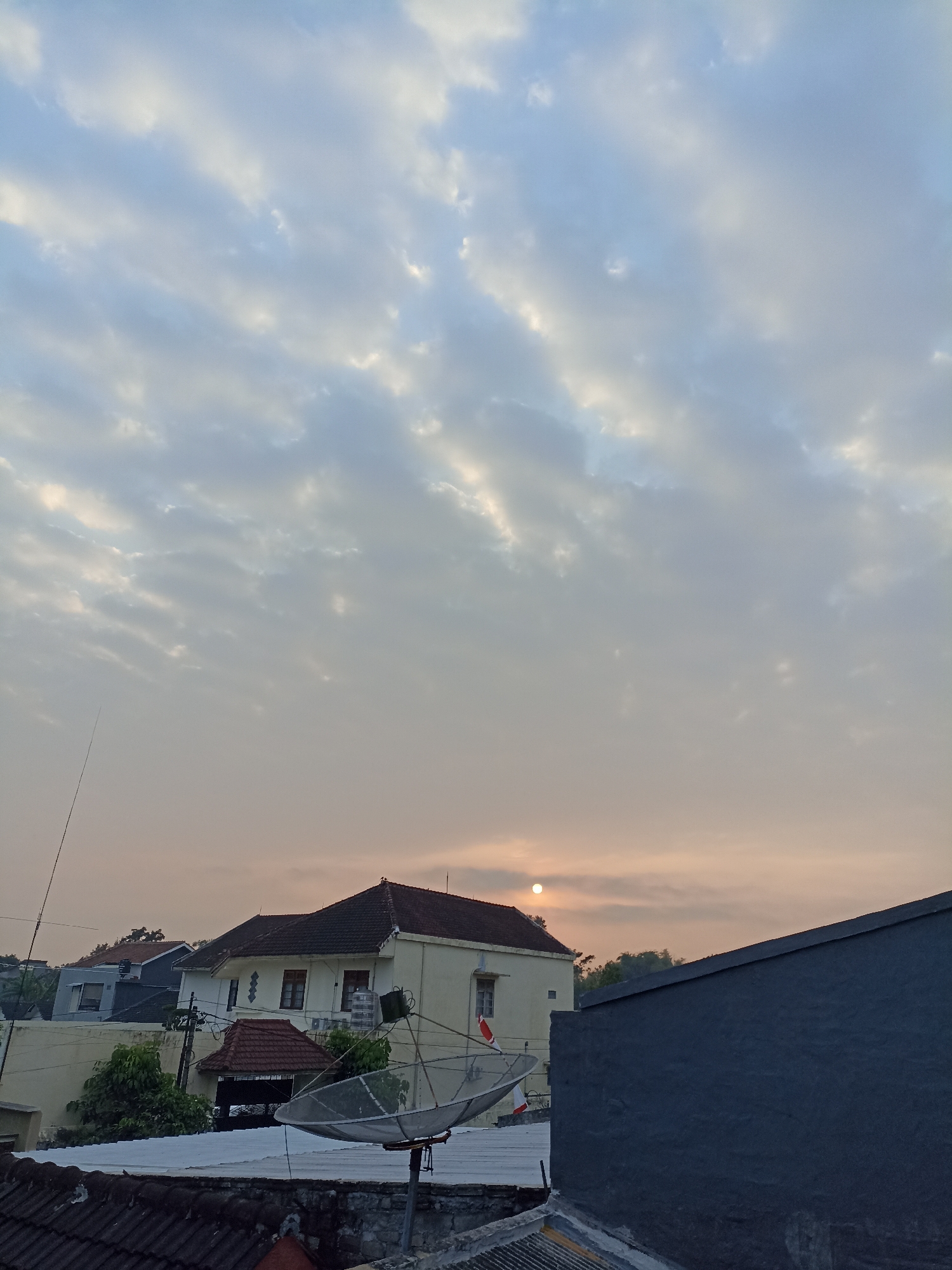 A wide shot captures a sky filled with textured clouds during what appears to be dawn or dusk. The sun, a small bright orb, is visible low in the sky on the left, partially obscured by clouds, casting a warm, orange-tinted light. In the foreground, the rooftops of several buildings are visible. A large, dark gray wall occupies the right side of the frame, extending into the foreground. To the left of this wall, a light-colored house with a red tile roof is prominent. A satellite dish is mounted on a stand in front of this house. A tall, thin antenna extends upwards from behind the house on the left. The overall scene suggests a residential area within the city of Sumberejo, Indonesia, with the soft, diffused light indicating either early morning or late evening. No people are visible in the image.
