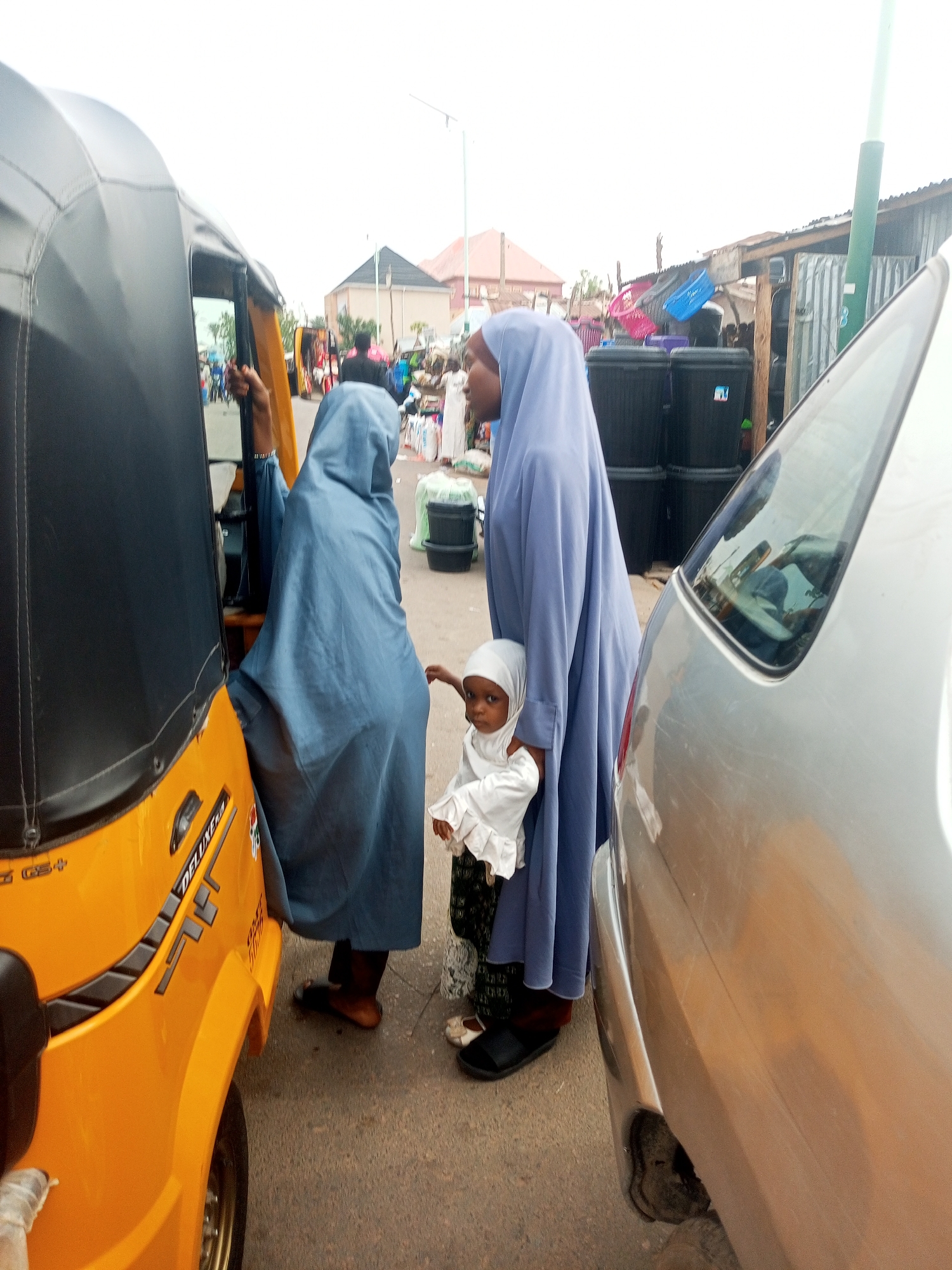 In Gombe, Nigeria, two women, both wearing hijab head coverings with long robes, are standing on a street next to a vehicle and a yellow tricycle. One woman holds a young child, who is also wearing a hijab. The environment includes buildings, black plastic bins, and other signs of commercial activity.
