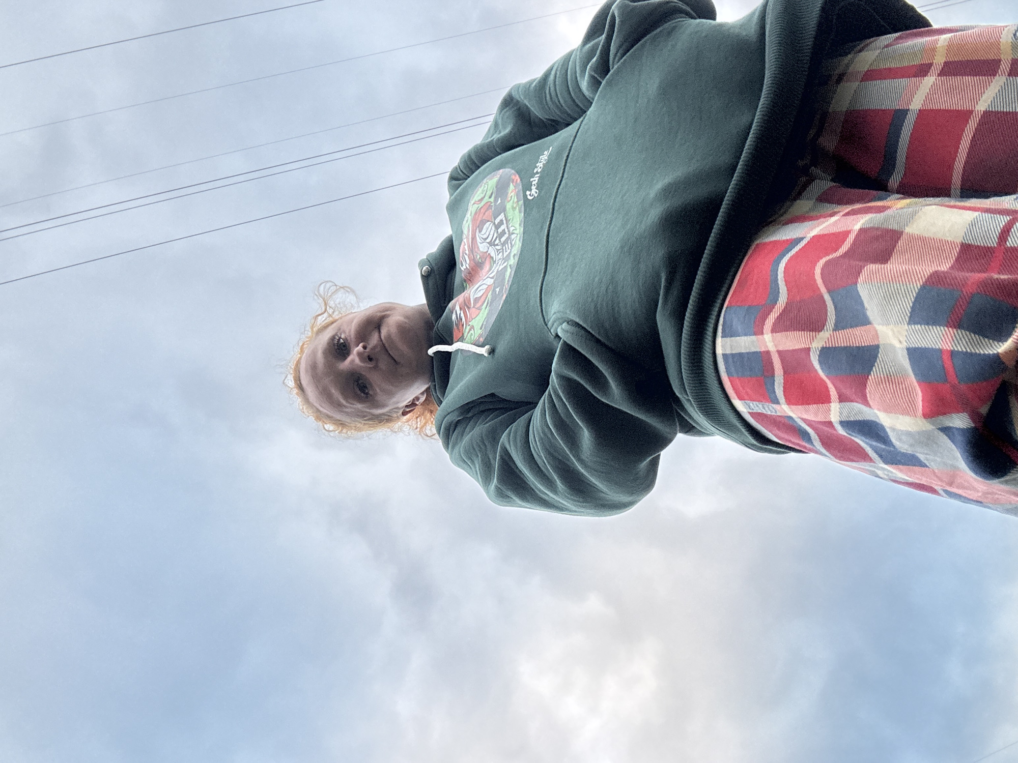 A low-angle, full-body shot of a person with curly red hair, wearing a dark green hooded sweatshirt with a graphic design and text that reads "Gesh Style" on the front, and red, blue, and white plaid pajama pants. The person is looking down at the camera with their hands in the pockets of the sweatshirt. The background is a cloudy sky with visible power lines stretching across it. The image was taken in Chaikovsky, Russia.