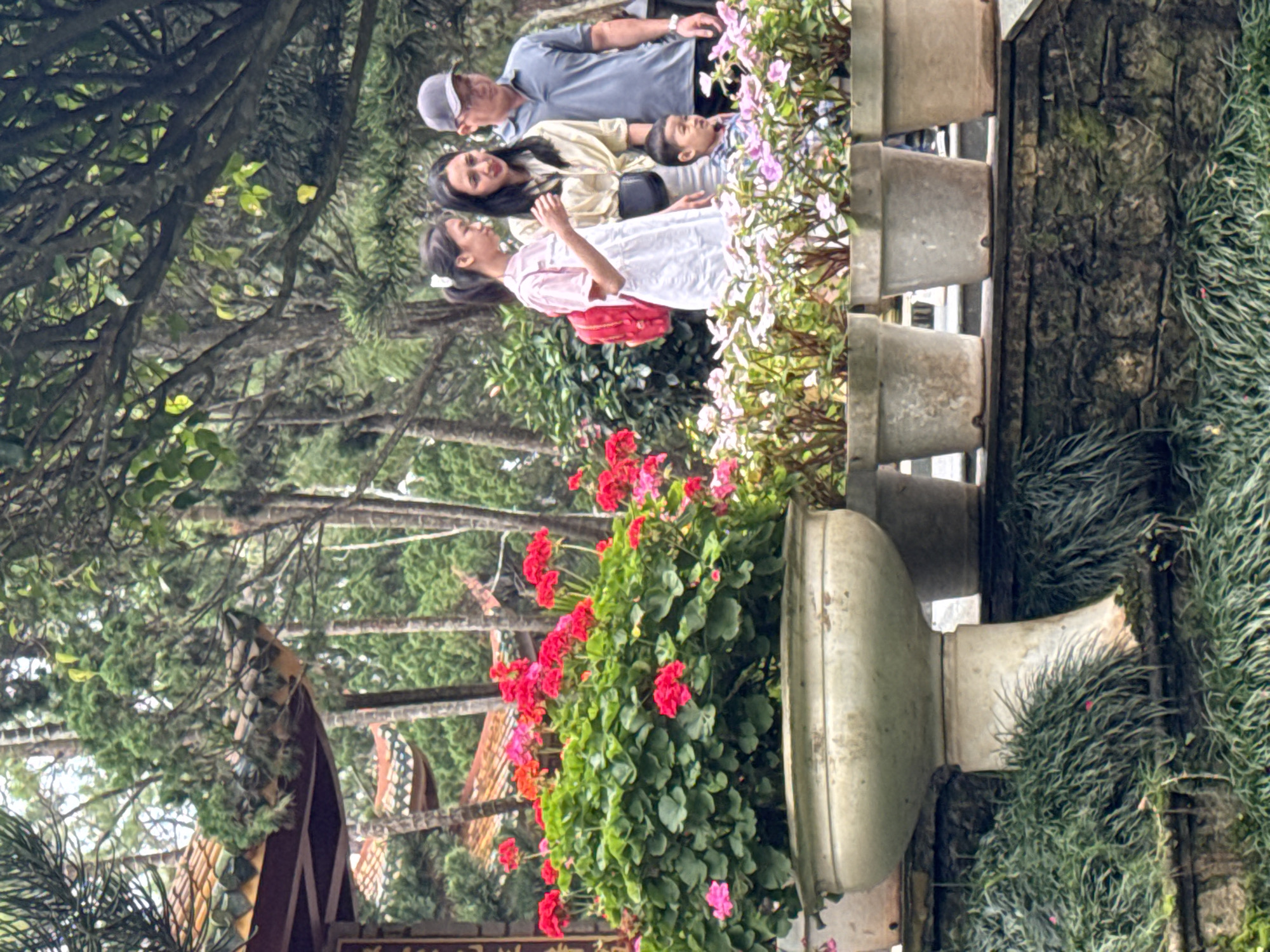 In Da Lat, Vietnam, a family is captured in a moment of leisure in what appears to be a botanical garden or park. The scene features lush greenery and vibrant flowers, with traditional Vietnamese architecture visible in the background. A man, a woman, and two children are the central subjects. The man, wearing a grey cap and a blue polo shirt, stands to the right. Beside him, a woman in a yellow shirt and white pants gazes to her left. A young girl with a ponytail, dressed in a pink and white outfit and carrying a red backpack, stands to the woman's left. A young boy, in a blue and white checkered shirt, is positioned in front of the adults, looking slightly upwards. The family is surrounded by potted plants, with a large white urn filled with red flowers dominating the foreground. The environment suggests a tranquil, well-maintained outdoor space with a hint of cultural significance.