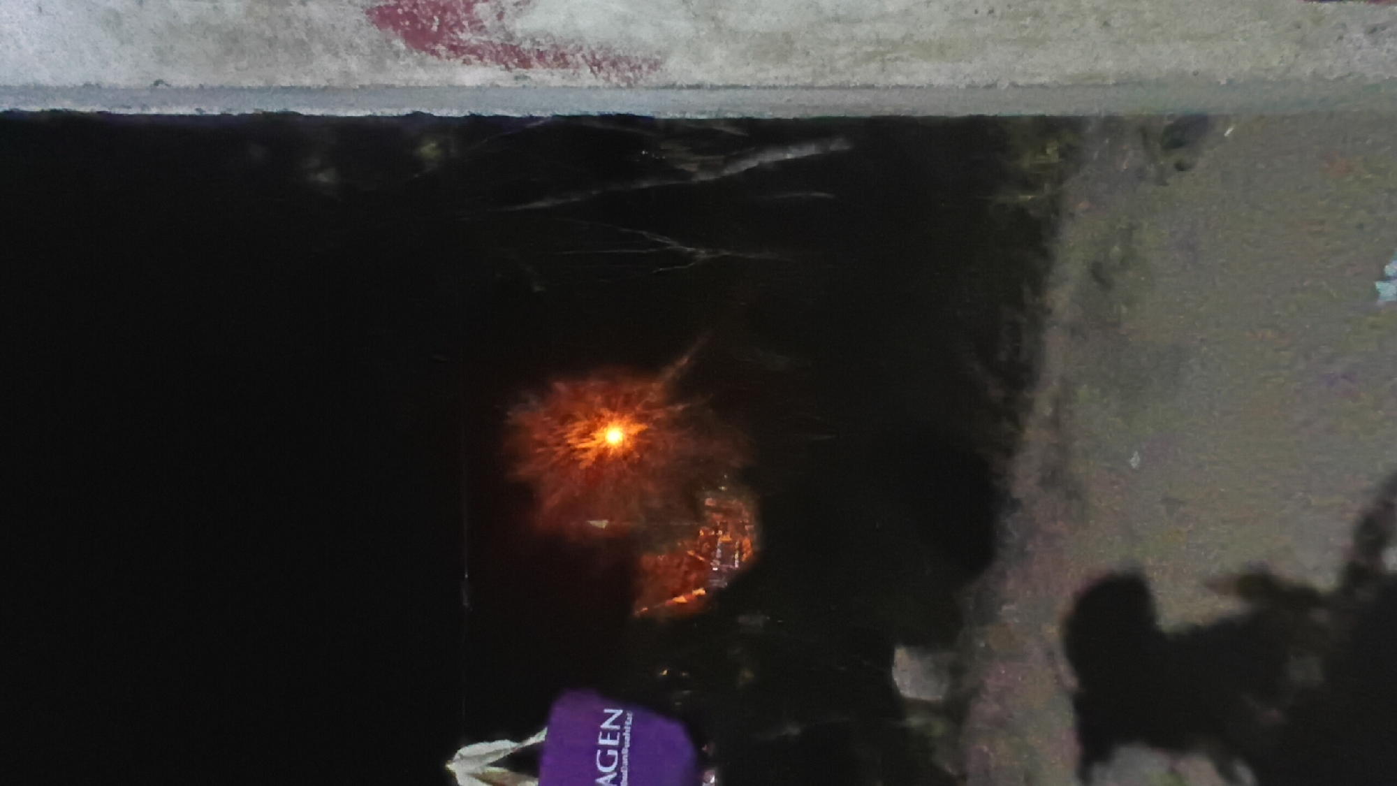 A photograph taken at night shows a blurry, indistinct scene with a bright light source in the middle distance, resembling an explosion or a very bright, diffused lamp. To the left, a portion of a purple bag with white lettering is visible. The foreground is dark and features a shadowy silhouette of a person or figure. A concrete pillar with some red markings is on the far right of the image. The background consists of dark, shadowy trees and foliage. The scene is likely outdoors, possibly in an urban or semi-urban area, given the presence of the pillar and the implied lighting. Without further context or clearer details, pinpointing the exact location within Prabumulih, Indonesia is not possible.