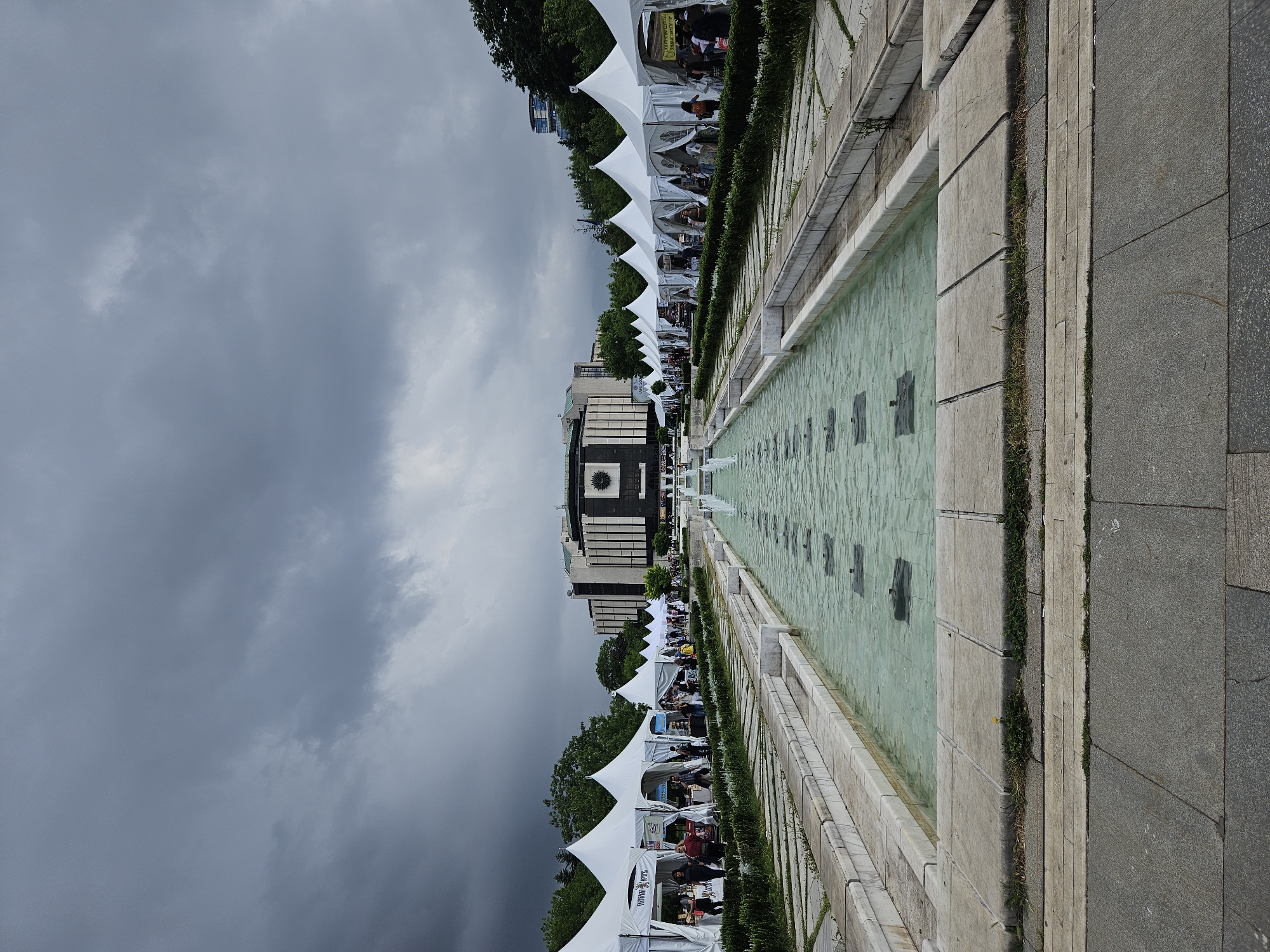 An outdoor scene in Sofia, Bulgaria, is captured under an overcast, cloudy sky, suggesting daytime. The image, originally rotated, depicts a long, narrow water feature or fountain running from the foreground towards a prominent building in the distance. The water, a light greenish-blue, contains several dark, rectangular shapes evenly spaced along its length, some of which appear to be indistinct symbols or letters.
To the left of the water feature, a long row of white, pointed event tents is set up, stretching into the background. Hedges line the path between the tents and the water. Numerous people are visible under and around these tents, some seated, others standing, indicating an active outdoor event, market, or fair. Behind the tents, lush green trees are visible, and further back, a large, modern-style building with a dark facade, multiple horizontal windows, and a distinctive clock face near its center stands prominently. This building is characteristic of the National Palace of Culture in Sofia.
To the right of the water feature, paved walkways extend, showing textures of concrete and granite slabs, with some small weeds growing in the cracks. The general atmosphere is busy and social. While some text is visible on banners under the tents and possibly in the water, it is largely illegible due to distance and resolution.
