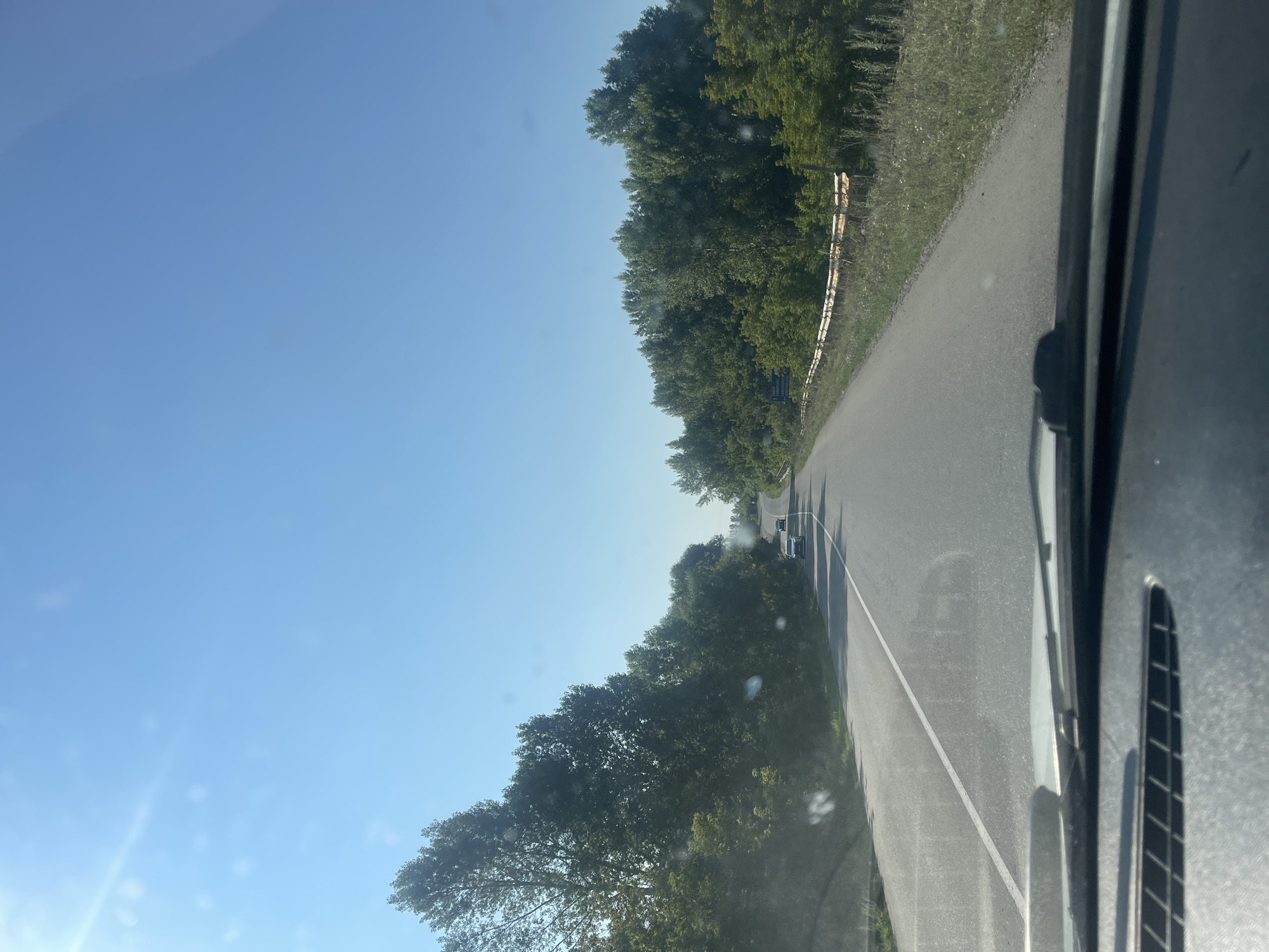 The image depicts a view from a vehicle driving on a paved road on a sunny day. The road is flanked by dense green trees on both sides, with a clear blue sky overhead. In the distance, two cars are visible on the road, moving in the same direction as the viewer. A guardrail runs along the right side of the road, separating it from a grassy verge and more trees. There are some blurry white spots on the windshield, possibly from water droplets or dust. The foreground shows the dashboard of the vehicle, including the windshield wipers. The scene appears to be a rural or semi-rural area, likely within the city of Samsonovka, Kazakhstan, given the provided context.