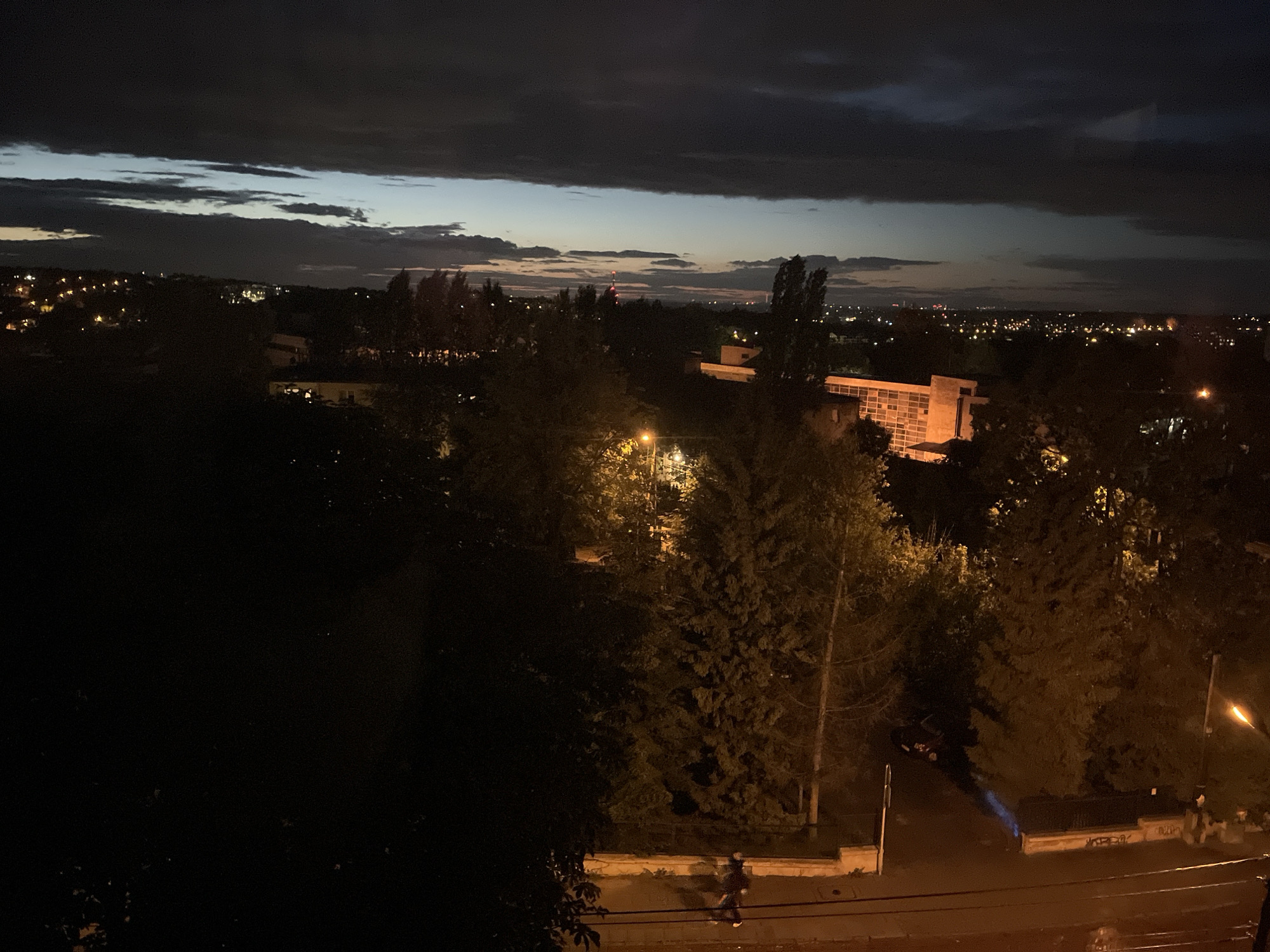 An elevated wide shot captures a dusky cityscape at night, with a focus on the transition from twilight to darkness. The sky is dominated by heavy, dark clouds with lighter, elongated bands of pale blue and yellow clouds near the horizon. Below the sky, the city unfolds with scattered lights from buildings and streetlamps. In the middle ground, a modern building with illuminated windows and a prominent rectangular structure stands out against the darker landscape. Trees, some silhouetted and others faintly lit by nearby lights, dot the scene. In the foreground, a street or pathway is visible, with a few dark silhouettes of people walking and a dark-colored car parked. A streetlamp casts a warm glow on the scene. The overall atmosphere is one of a peaceful, urban evening.