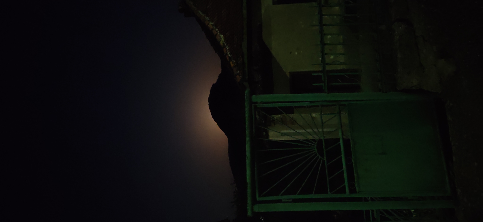 The image is a nighttime outdoor photograph taken in the city of Gara Lakatnik, Bulgaria. In the foreground, a green metal gate with a sunburst design is visible. Behind the gate, a dark silhouette of a building with a tiled roof is present. Further in the background, a rounded hill or mountain is silhouetted against a dark sky. A faint glow, possibly from distant lights or the moon, is visible behind the crest of the hill. The overall scene is dark, with the primary illumination coming from an unseen source that casts a green hue on the gate.