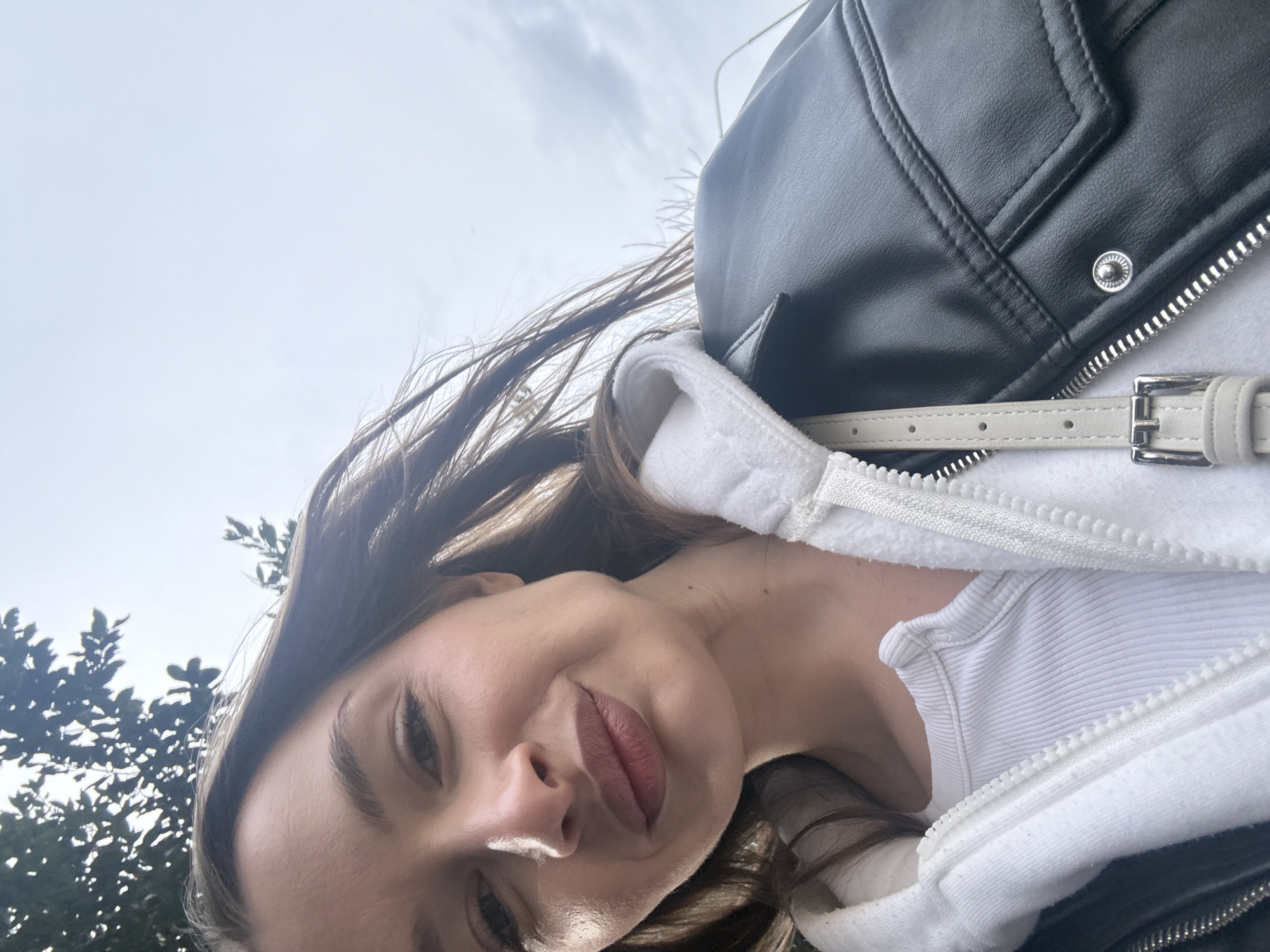 A selfie shot angled upward shows a woman with fair skin and brown hair. She is wearing a white hoodie partially zipped up under a black leather jacket with a silver zipper and silver hardware on the collar. A thin, off-white belt strap with a silver buckle is visible over her shoulder. She wears a white ribbed tank top under the hoodie. The background features a light sky with clouds and green tree foliage in the lower left corner. The specific location in Budva, Montenegro, cannot be determined from the given visual information.