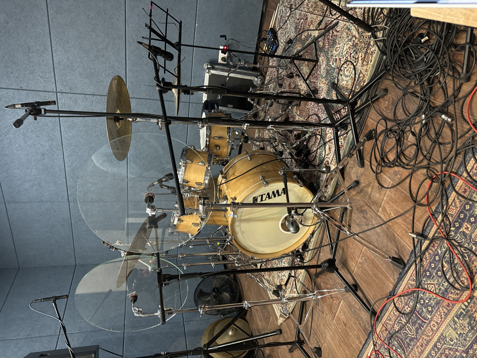 The image displays an unoccupied, fully-miked drum kit positioned within a sound-treated recording studio located in Moscow, Russia. The central object is a light-colored wooden five-piece Tama drum kit, comprising a bass drum (bearing the "TAMA" logo), a snare drum, and three tom-toms. Accompanying the drums are hi-hats, a crash cymbal, and a ride cymbal. Two large, transparent acrylic drum shields are erected in front of the kit.
Multiple microphones are strategically placed on boom stands and clamps around the drum kit, targeting individual drums (snare, tom-toms, bass drum interior), hi-hats, and overheads for comprehensive audio capture.
The studio environment features walls covered with blue-gray acoustic panels and a floor composed of light-brown wooden planks partially covered by an ornate, patterned rug with red, blue, and cream hues situated beneath the drum kit.
Additional visible objects include a black floor fan positioned to the left rear of the kit, a stack of cymbals leaning against the wall further left, and a black music stand to the right. Numerous audio cables, primarily black with some red, are coiled and sprawled across the rug and wooden floor. In the background to the right, a silver equipment case, a power strip, and a small blue/black stompbox-style pedal are discernible. No individuals are present in the scene.