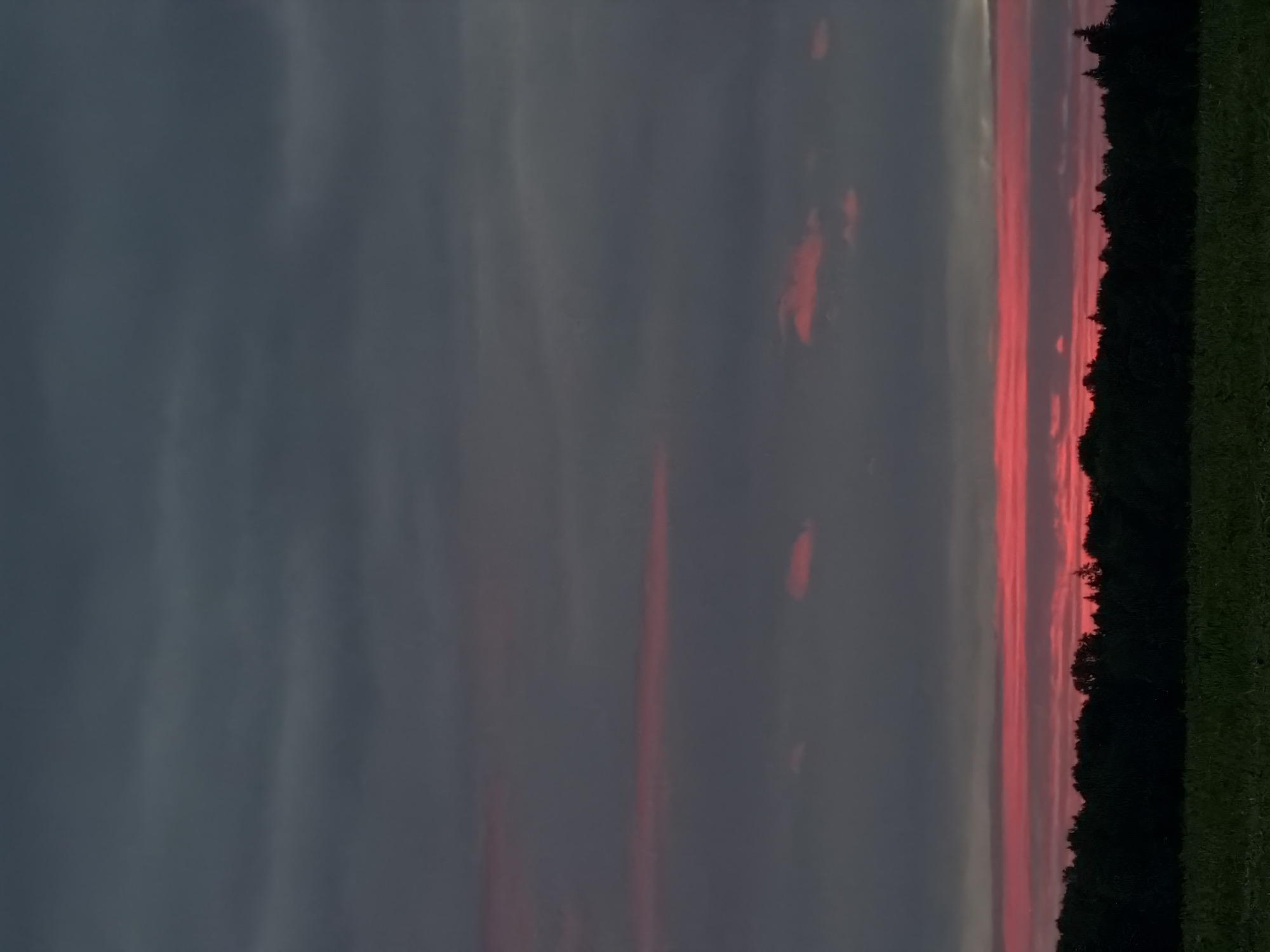 This vertical outdoor photograph depicts a landscape in Kechur, Russia, captured during twilight or sunset. The lower approximately 15% of the frame features a dark green field or grassy area. Immediately above the field, a dense, dark silhouette of a forest or treeline stretches horizontally across the image. The sky occupies the majority of the upper frame. The top portion of the sky is filled with layered, dark grey clouds. Below these dark clouds, a prominent, elongated band of intense red and pink clouds extends horizontally across the horizon, directly above the treeline. Additionally, scattered, smaller patches of red and pink are visible within the darker grey clouds in the middle section of the sky. No individuals, animals, or specific man-made structures are discernible within the scene.