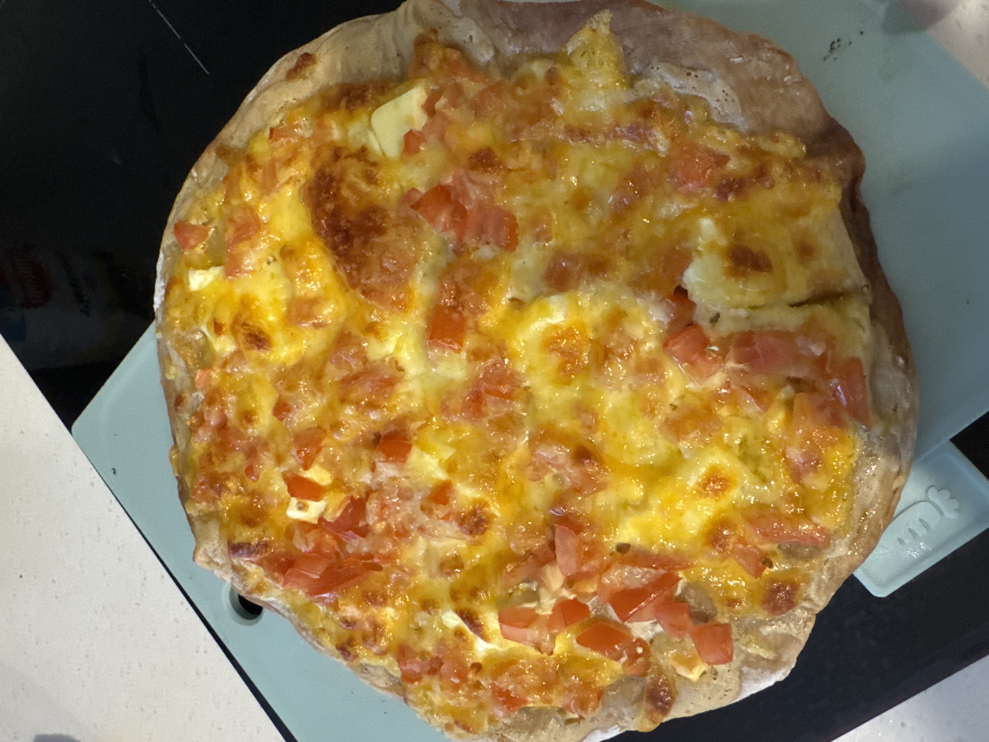 A close-up, high-angle shot shows a round, homemade pizza on a light blue cutting board, which is resting on a black surface. The pizza is topped with melted, golden-brown cheese and diced red tomatoes. The crust is a light brown color and appears slightly raised. A small, light blue kitchen utensil with a bunny design is visible to the left of the pizza, partially under the cutting board. The black surface beneath the cutting board has some faint white lines.