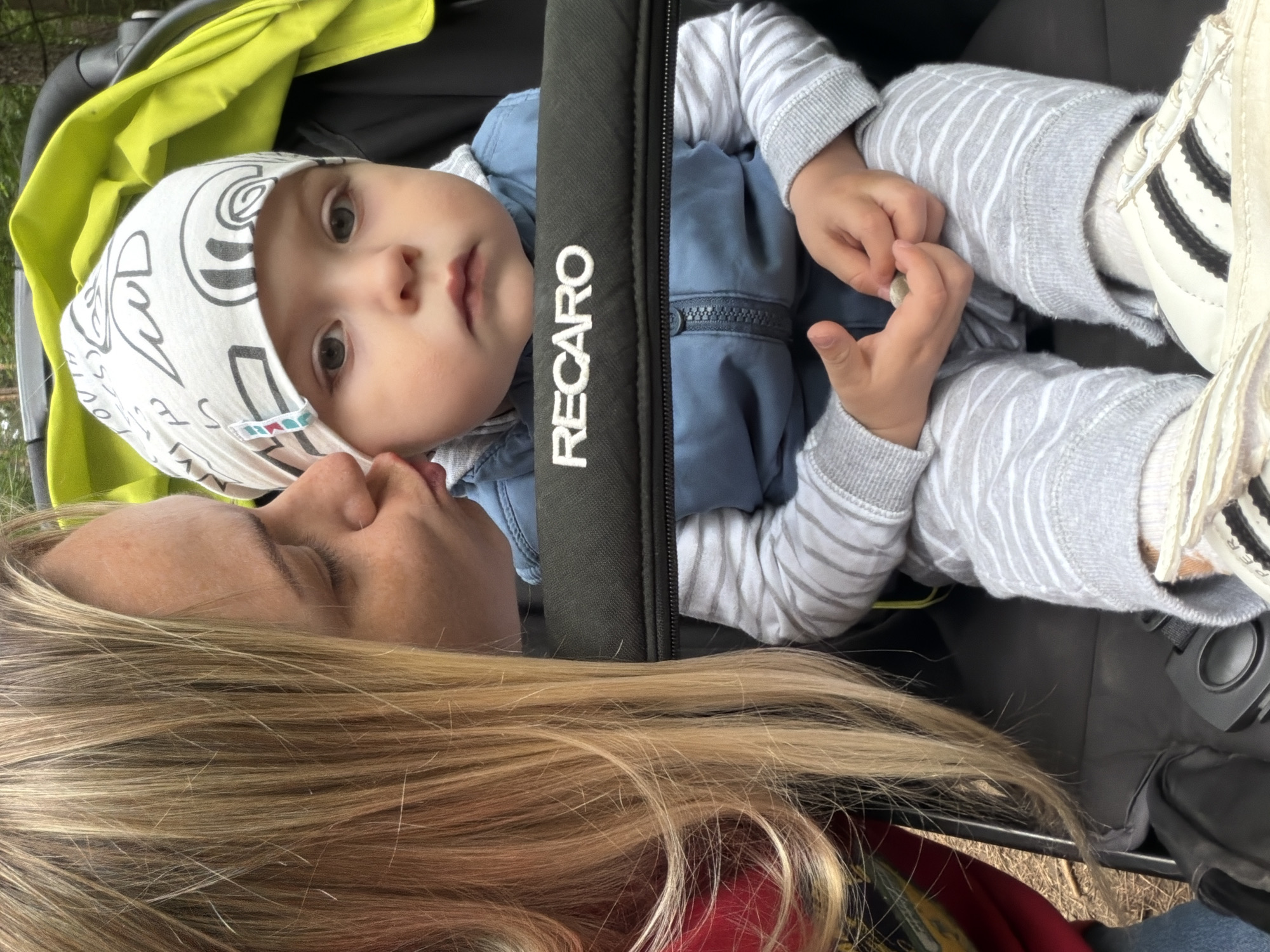 In Votkinsk, Russia, a blonde woman leans in to kiss a baby seated in a Recaro stroller. The woman is wearing a red top, and the baby is dressed in a blue vest over a striped shirt and pants, with a white patterned hat. The stroller's bright yellow canopy is visible behind the baby. The scene appears to be outdoors, possibly in a park or wooded area, given the visible foliage in the background and the dry leaves on the ground.