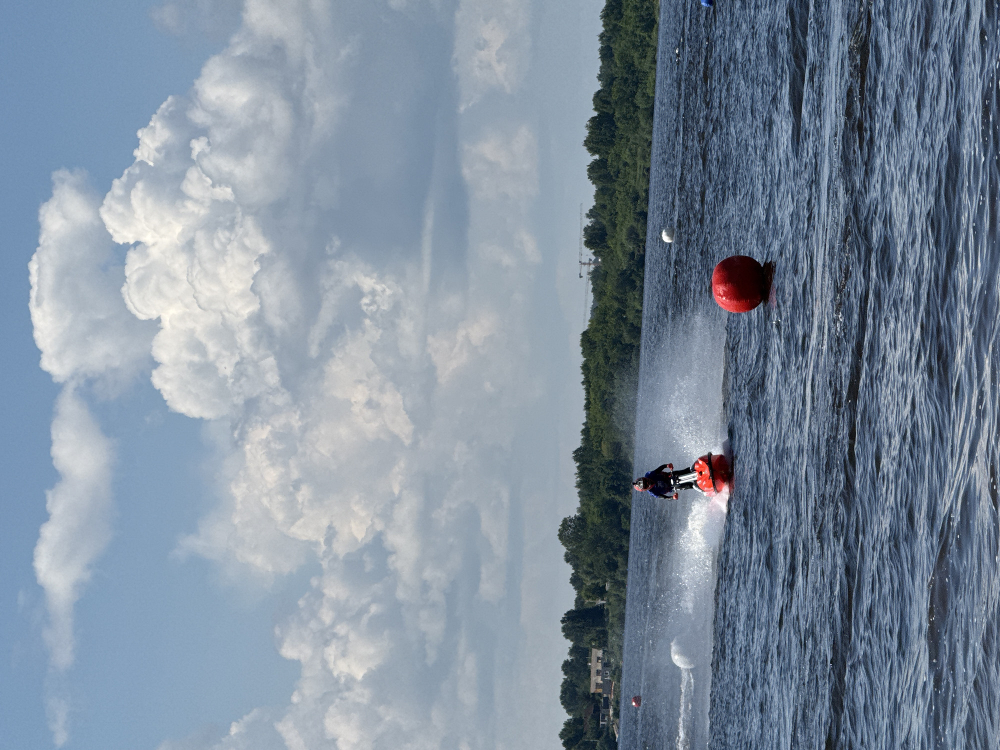 A person is riding a red jet ski on a body of water, creating a spray behind them. They are wearing a helmet and a dark blue and red racing suit. In the background, there is a line of trees and buildings on the shore, with a cloudy blue sky above. There are several buoys in the water, including a large red one in the foreground and a white one to the left of the jet ski. In the distance, construction cranes are visible above the trees. This scene is likely taking place on the Kama River in the city of Perm, Russia, during a jet ski event or recreational activity.