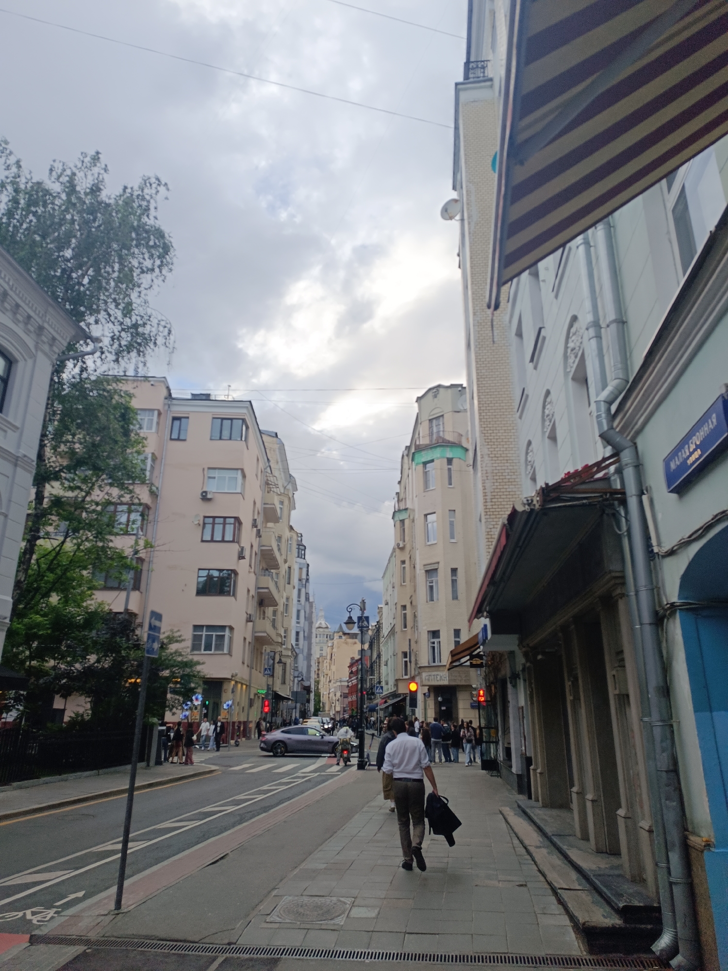 An eye-level, wide shot captures a street scene in an urban environment under a cloudy sky. On the right side of the frame, a building features a striped awning and a blue street sign reading "МАЛАЯ БРОННАЯ". The building's facade is light blue and adorned with decorative elements. To the left, a street with cars and pedestrians unfolds. A person in a white shirt and brown pants walks away from the camera, carrying a black bag. Another person is on a motorcycle in the distance. The street is lined with trees and multi-story buildings. The sky is overcast with grey clouds.
