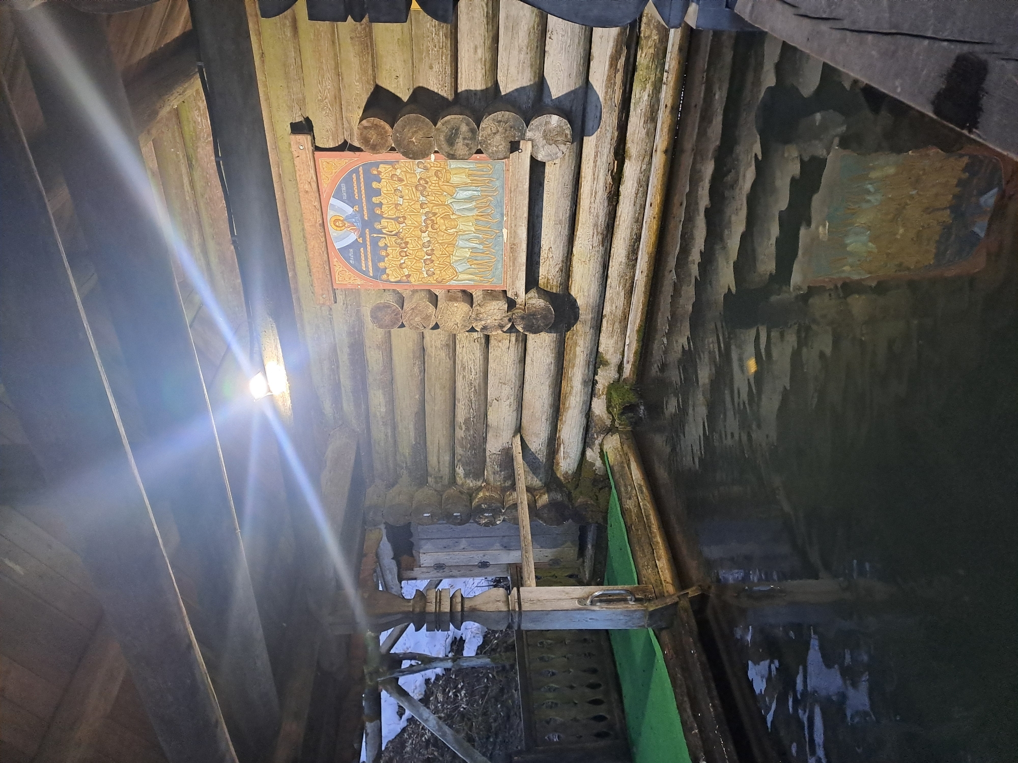 The image depicts the interior of a wooden structure, likely a wellspring or small building, located near Vzglyadnevo, Russia. A religious icon is affixed to the log wall, held in place by wooden posts. Water is present in the structure, reflecting the surrounding wooden walls and roof. A ladder or steps leads down into the water. Debris, including leaves and snow, are visible near the base of the steps. A green covering is positioned near the water's edge. Artificial light illuminates the scene from the left side.