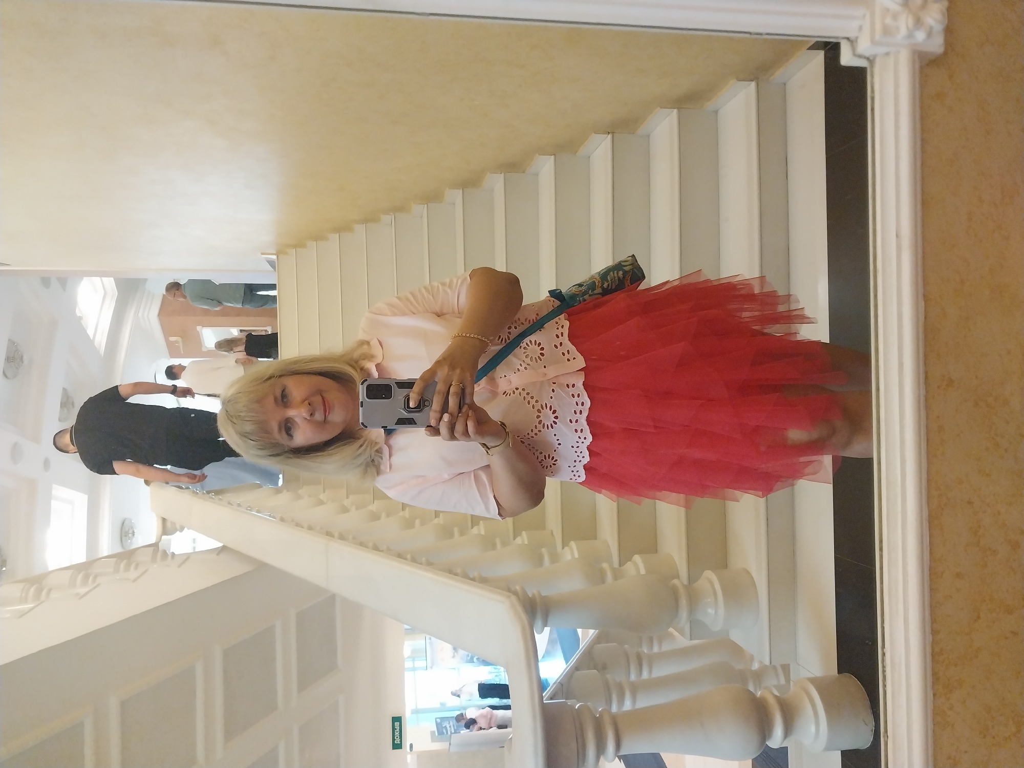 A blonde woman is taking a mirror selfie on a white staircase. She is wearing a light pink collared shirt with eyelet detailing around the hem and a voluminous red tulle skirt. She holds a smartphone in her right hand, and a patterned crossbody bag hangs from her left shoulder. In the reflection, several other people are visible on the stairs and in the background, which appears to be an indoor public space, possibly a museum or cultural center. One man in a black t-shirt is ascending the stairs in front of her. The location is identified as the Orenburg State Regional Museum of Local Lore, Orenburg, Russia.