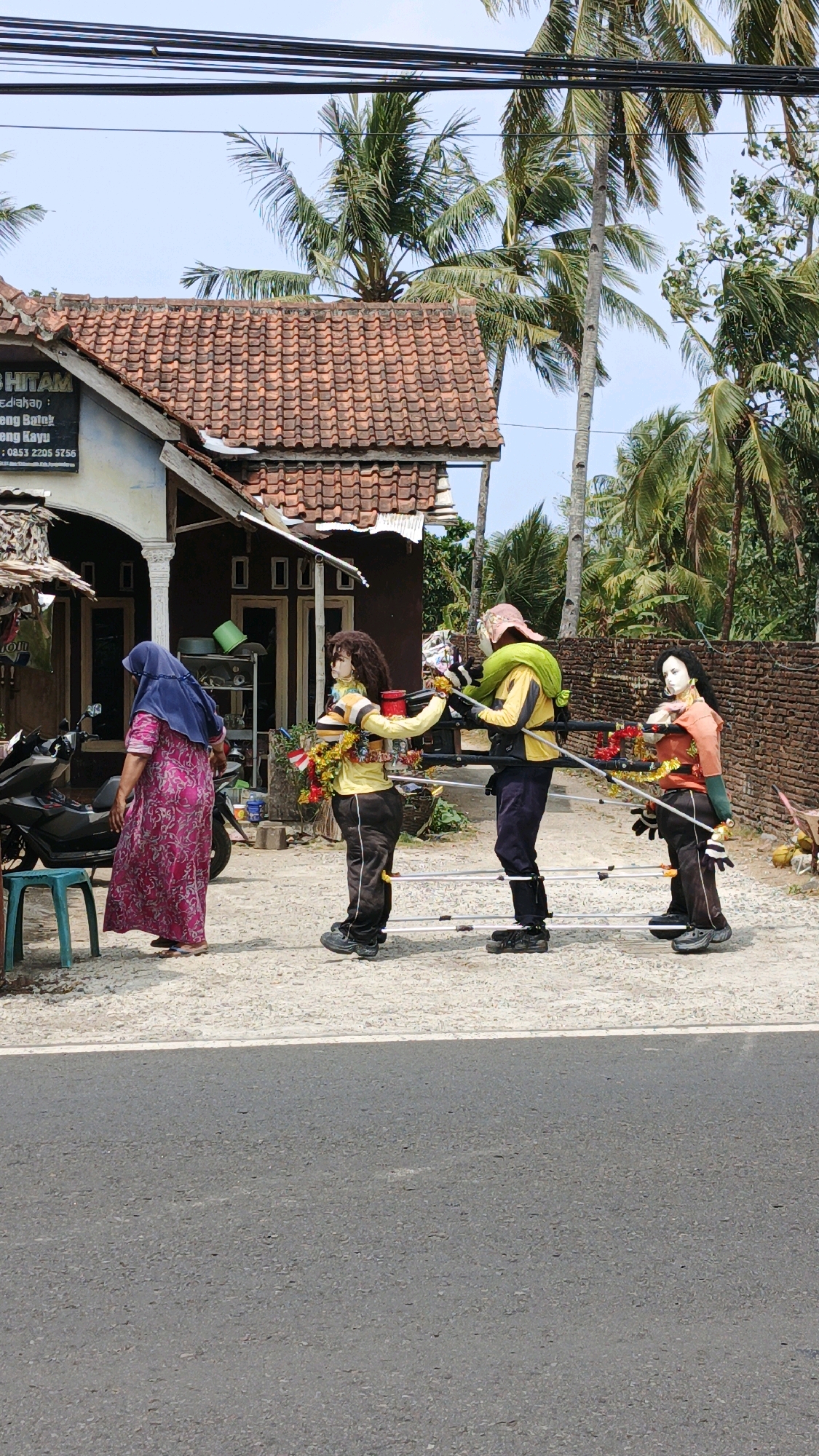 This image, captured in Sukaresik, Indonesia, depicts an outdoor scene with a focus on a parade or procession of three figures. The figures appear to be mannequins or scarecrows dressed in clothing and decorated with garlands of flowers and small Indonesian flags. The first mannequin, on the far left, is partially obscured by a woman wearing a pink patterned dress and a blue headscarf. This woman is looking away from the camera, towards the figures. The other two mannequins are being carried on a makeshift wooden structure, supported by two individuals. The mannequin in the middle is wearing a yellow shirt and dark pants, and the one on the right is in an orange shirt and dark pants. Both carriers are also wearing dark pants and their faces are covered with masks. They are walking in a line on a gravel path adjacent to a paved road. Behind the figures is a building with a red tiled roof and a dark brown facade, and further in the background, palm trees and a brick wall are visible. The lighting suggests it is a sunny day with clear skies. A parked motorcycle is also present near the woman.