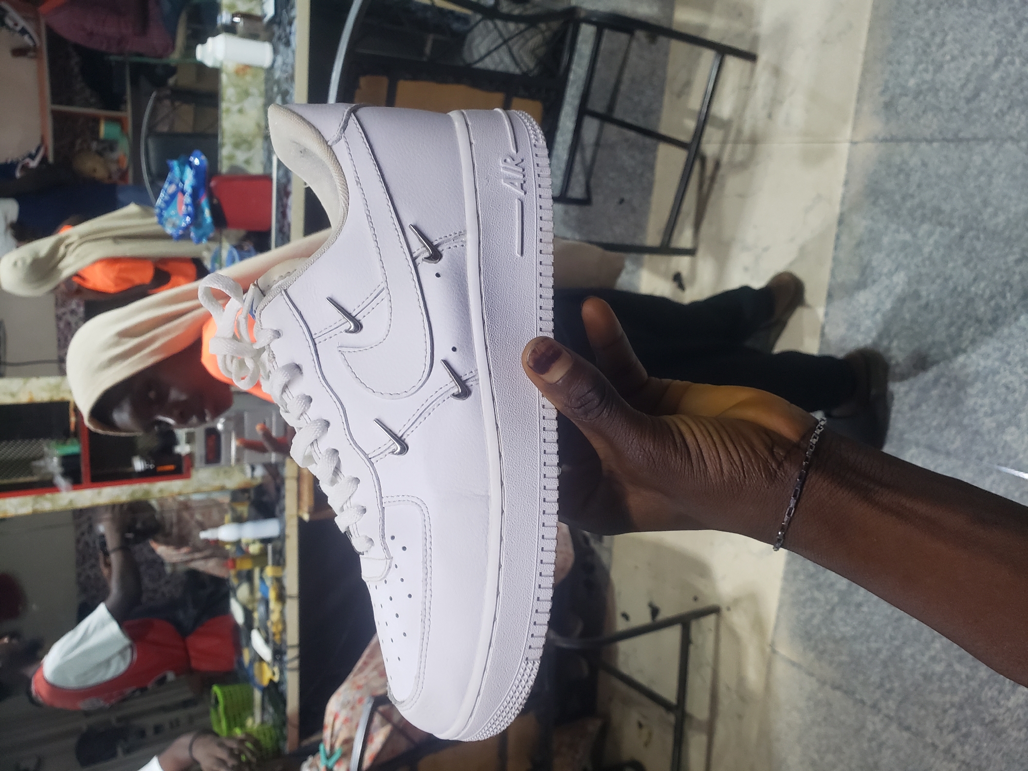 In a retail environment, possibly a barber shop or boutique, a person's hand is holding up a white Nike Air Force 1 sneaker. The sneaker is clean and white with subtle metallic Nike swooshes. In the blurred background, several individuals are visible, including a person in a red vest and a person wearing a headscarf. The environment appears to be indoors, with shelves and furniture visible. The flooring is tiled.