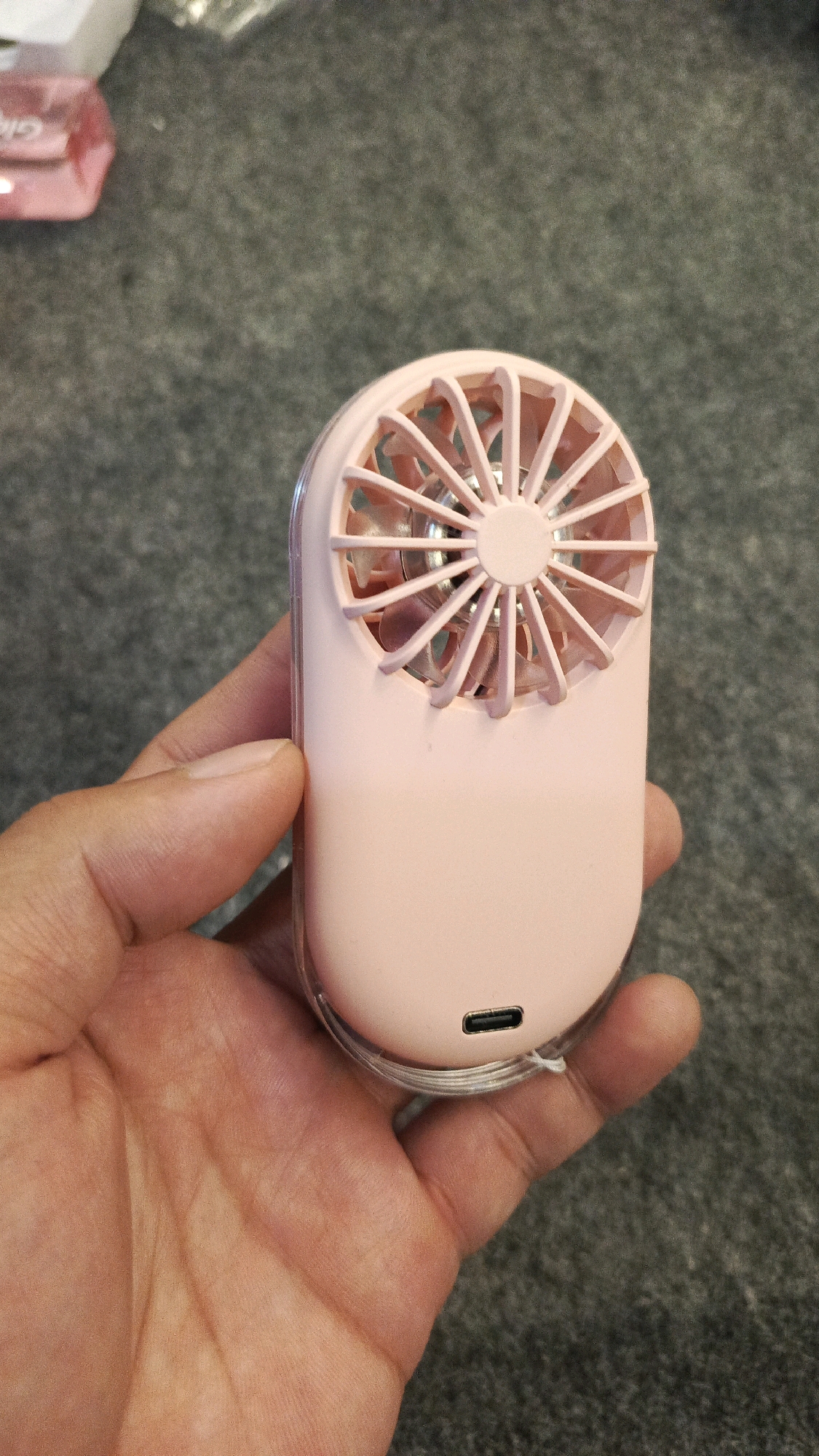 A person's hand holds a pink portable fan with a clear plastic case. The fan has a circular grill with radial spokes and a central circular element. A USB-C port is visible on the bottom of the fan. The background is a grey, textured surface.