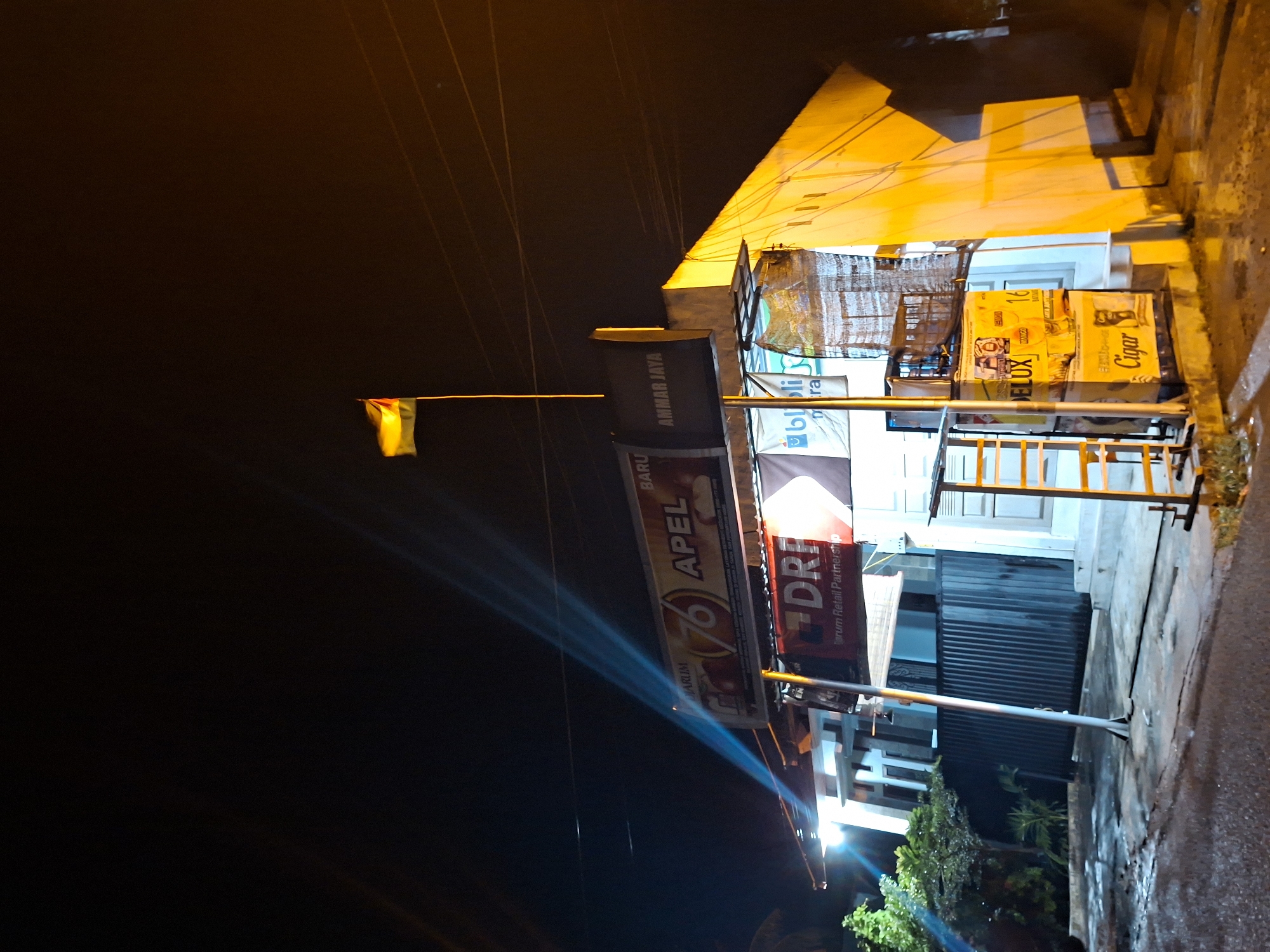 Captured at night in Lhokseumawe, Indonesia, the photograph showcases a brightly lit shop front. The street is dimly lit, but the store features numerous illuminated signs: "APEL," "DRF," and "AMMAR JAYA," among others. Various advertising boxes are stacked near the entrance. Overhead electrical wires stretch across the dark sky, with a small, horizontally extended flag visible towards the upper left. The environment appears to be wet, with reflections on the street surface, and some greenery is visible near the shop front.