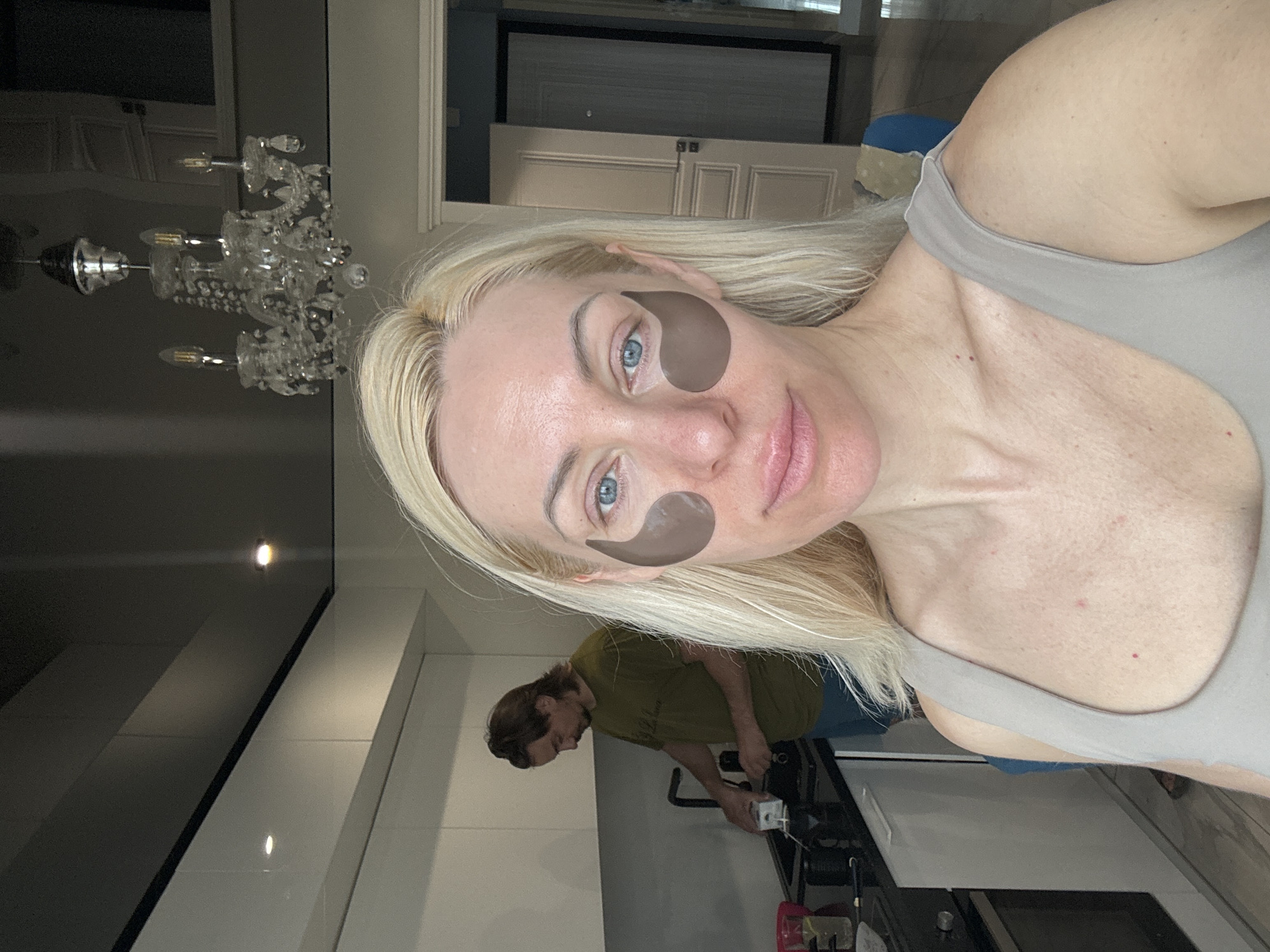 A selfie of a woman with light blonde hair, light skin, and blue eyes wearing brown under-eye masks. She is wearing a light gray tank top. A man in a dark green t-shirt and dark pants is in the background near a white kitchen counter and black stove. A decorative chandelier is also visible in the background. The location is Unknown, Unknown.