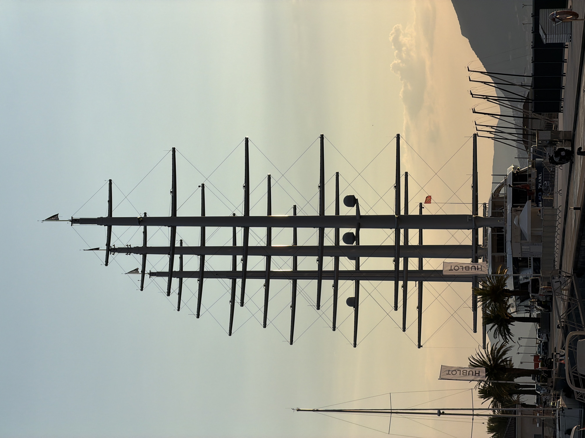 The image captures a large sailing yacht, the "Black Pearl," docked in Tivat, Montenegro, at sunset. The yacht dominates the frame with its towering masts and numerous horizontal spars, silhouetted against a gradient sky transitioning from pale blue at the top to a warm yellow-orange near the horizon. Several flags, including one with a skull and crossbones emblem and a red flag, are visible at the mastheads.
In the foreground, a waterfront promenade lines the marina. To the left, another sailboat with a tall mast is docked. Palm trees and other vegetation are present along the waterfront. Several vehicles are parked behind the promenade. To the right, a series of smaller masts, likely from other boats, are clustered together. In the background, a mountainous landscape is visible, softened by the twilight. The overall scene conveys a sense of tranquility and luxury associated with the marina.