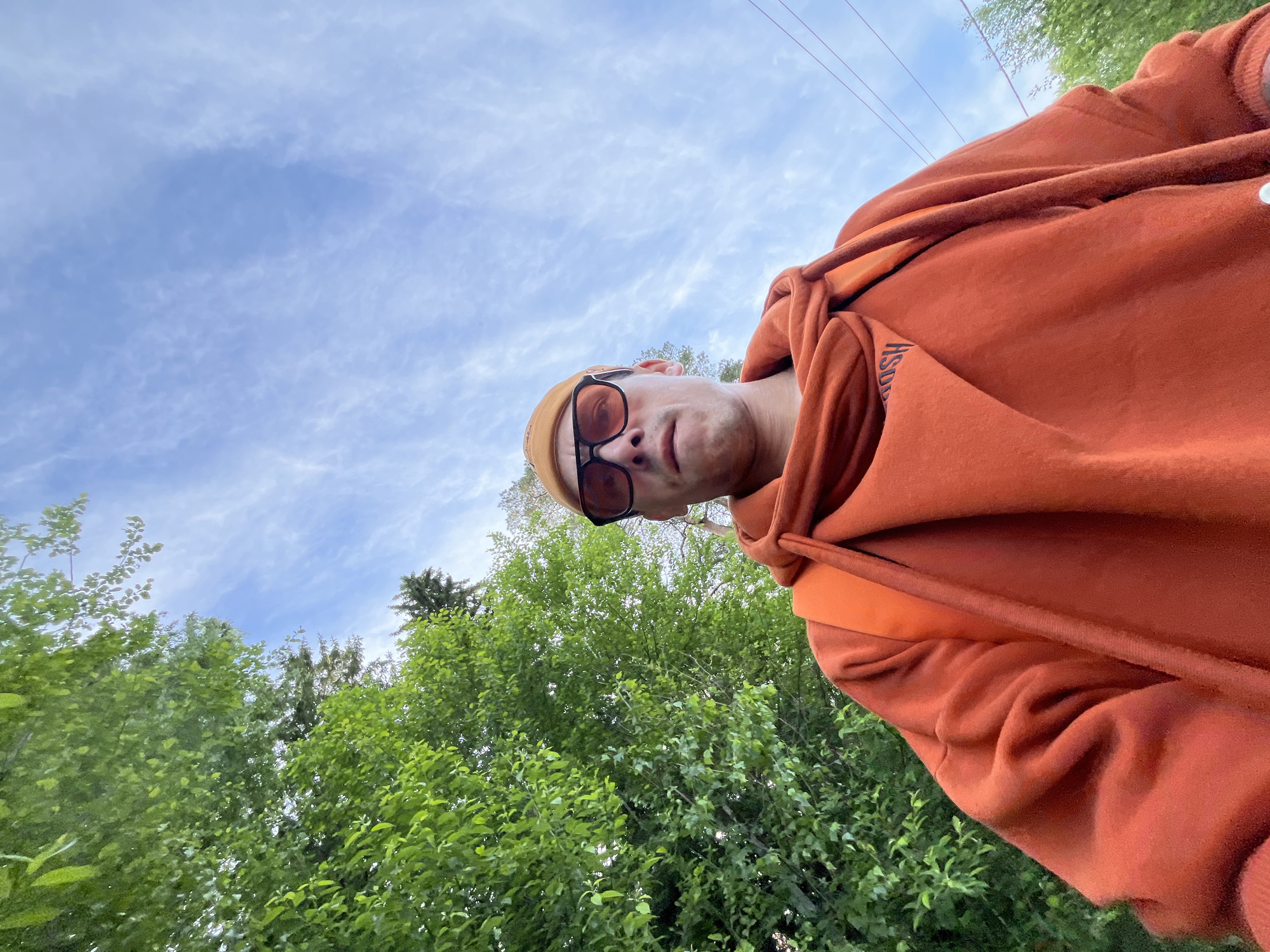 The image captures a low-angle selfie of a male individual outdoors in Chaikovsky, Russia. The subject is looking directly at the camera with a neutral expression. He wears a prominent dark orange or rust-colored hooded sweatshirt, which features dark, partially obscured text on the left chest area, possibly reading "HSDU". He also sports dark-tinted sunglasses and a light brown or beige beanie/cap.
The perspective is from below, with the subject's head slightly tilted, allowing a large portion of the upper frame to be filled by the sky. The sky is bright blue with scattered white, wispy clouds, suggesting a clear to partly cloudy daytime. In the lower background, lush green foliage from numerous deciduous trees dominates, with some darker evergreen trees visible further back. Faint power lines stretch across the upper mid-ground, cutting across the sky. The overall scene conveys a casual outdoor moment on a bright day.