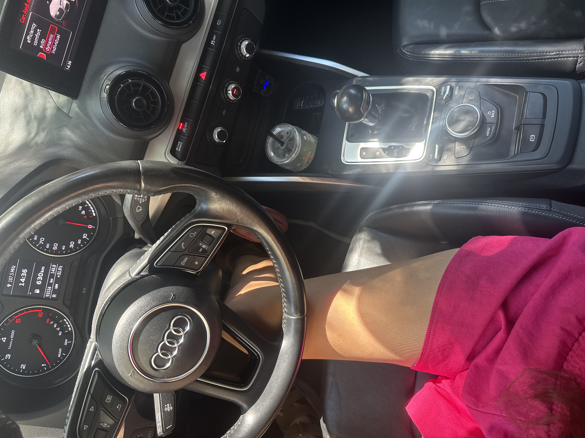 The image depicts the interior of an Audi vehicle, specifically the dashboard and center console area, viewed from the perspective of the driver or front passenger. The steering wheel is visible, bearing the Audi logo. The instrument cluster shows a digital display with time, speed, and distance readings. The center console includes the gear shifter, climate control buttons, and an infotainment system screen displaying drive select modes: "efficiency," "comfort," "auto," "dynamic," and "individual." A cup with a straw is placed in a cupholder. A person's leg, wearing sheer hosiery and a bright pink garment, is positioned in the foreground, obscuring a portion of the driver's seat and the lower part of the center console. The lighting suggests a daytime scene, with sunlight casting shadows within the car. There is no identifiable information within the image that would place this scene specifically in Plovdiv, Bulgaria.