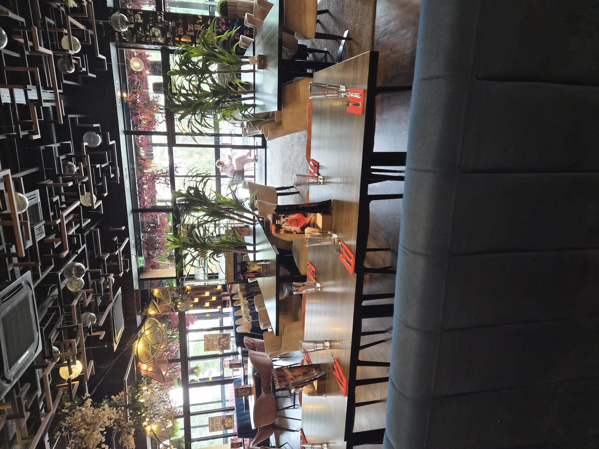 The image depicts the interior of a restaurant in Sofia, Bulgaria. The camera is oriented along a row of tables, set with glasses and red napkins, stretching from the foreground to the windows at the far end. Plants in pots are arranged along the tables near the windows. Blue booth seating is visible along one side of the tables. The ceiling is decorated with a geometric wooden structure with interspersed light fixtures. Through the windows, glimpses of greenery and an individual sitting are visible. The lighting is warm and ambient.