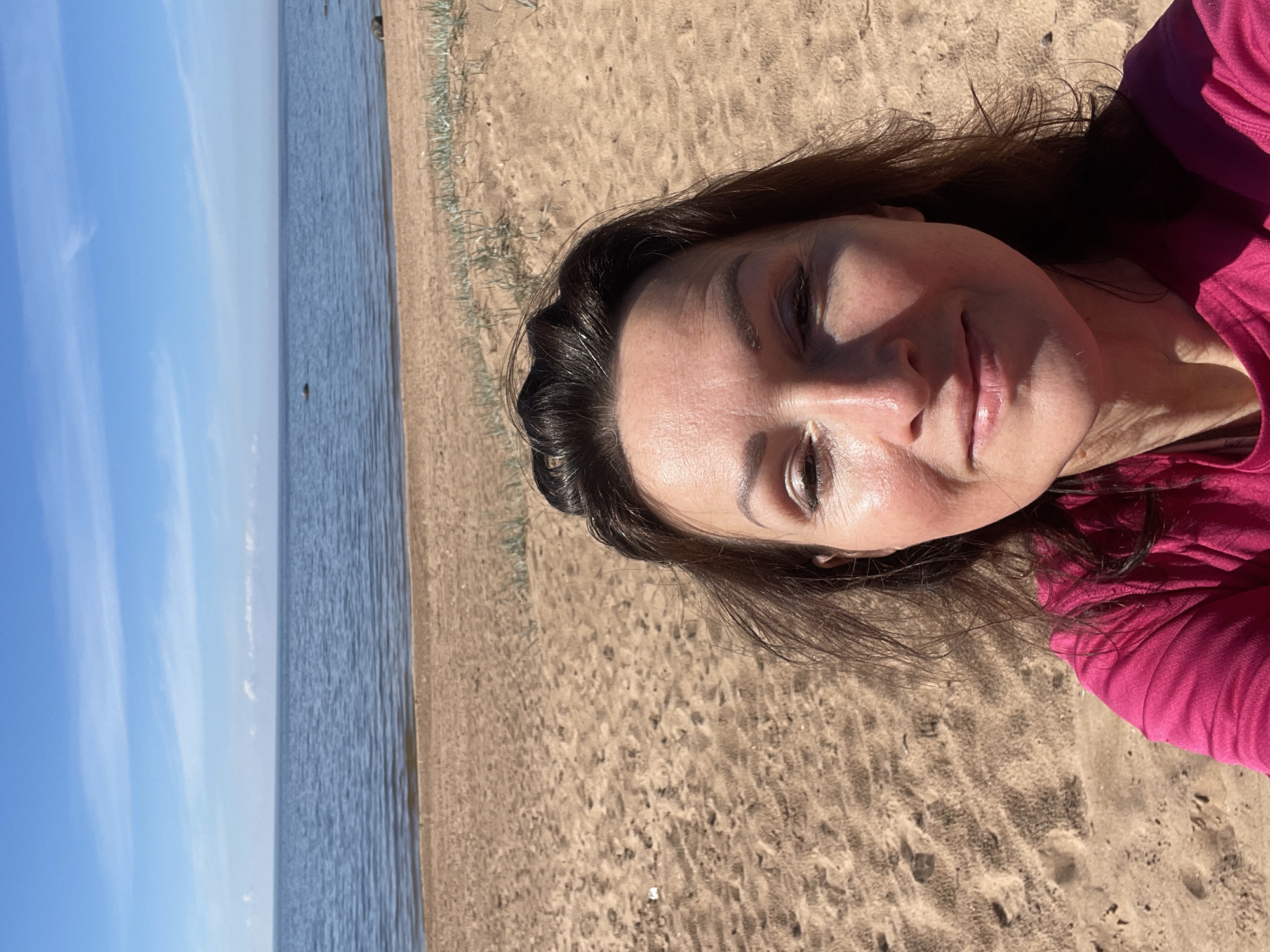 An outdoor, eye-level selfie-style photograph depicts a woman on a sandy beach under a clear blue sky in Zelenaya Roshcha, Russia. The woman, positioned in the lower half of the frame, has medium-brown hair, a slight smile, and her eyes are partially closed, with bright sunlight casting shadows on the right side of her face. She is wearing a magenta short-sleeved top. The immediate foreground and mid-ground consist of light brown sand with visible footprints and sparse green vegetation. A calm body of blue-grey water, likely the Gulf of Finland, occupies the central portion of the background, with several dark rocks visible partially submerged. A flat horizon separates the water from a bright blue sky with faint, wispy white clouds.