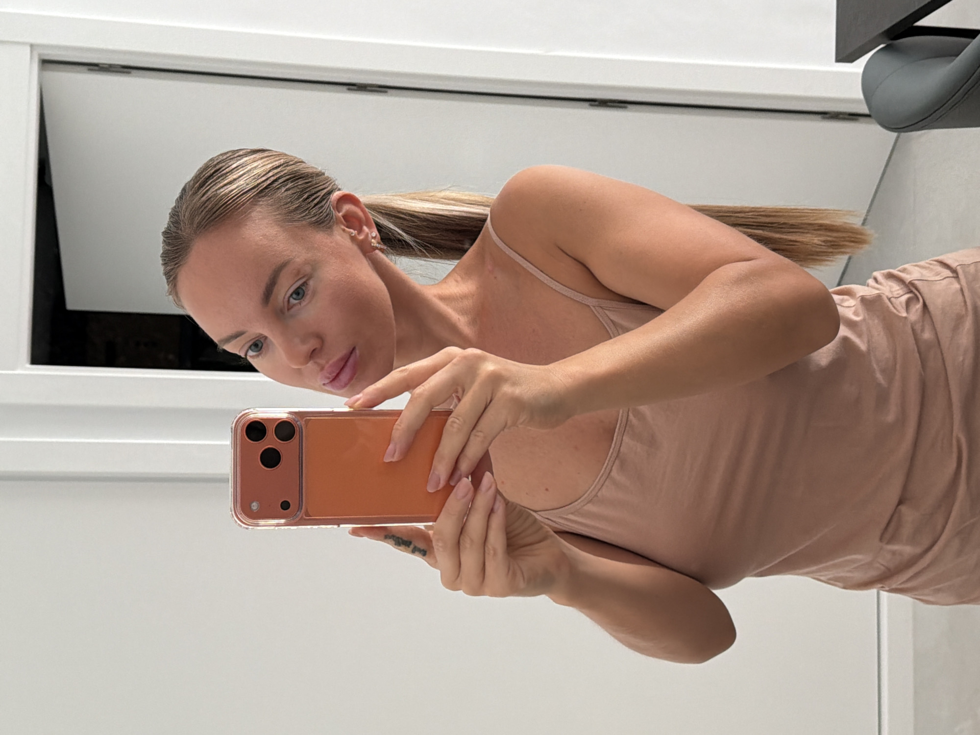 A young woman with blonde hair pulled back in a ponytail and wearing a pink tank top is taking a selfie in a mirror. She is holding an orange phone in her hands. The background is a white wall with a closed door to her right. A table and a grey chair are visible in the lower right corner of the image. The image is lit by natural light and appears to have been taken indoors. The location is identified as Finestrat, Spain.