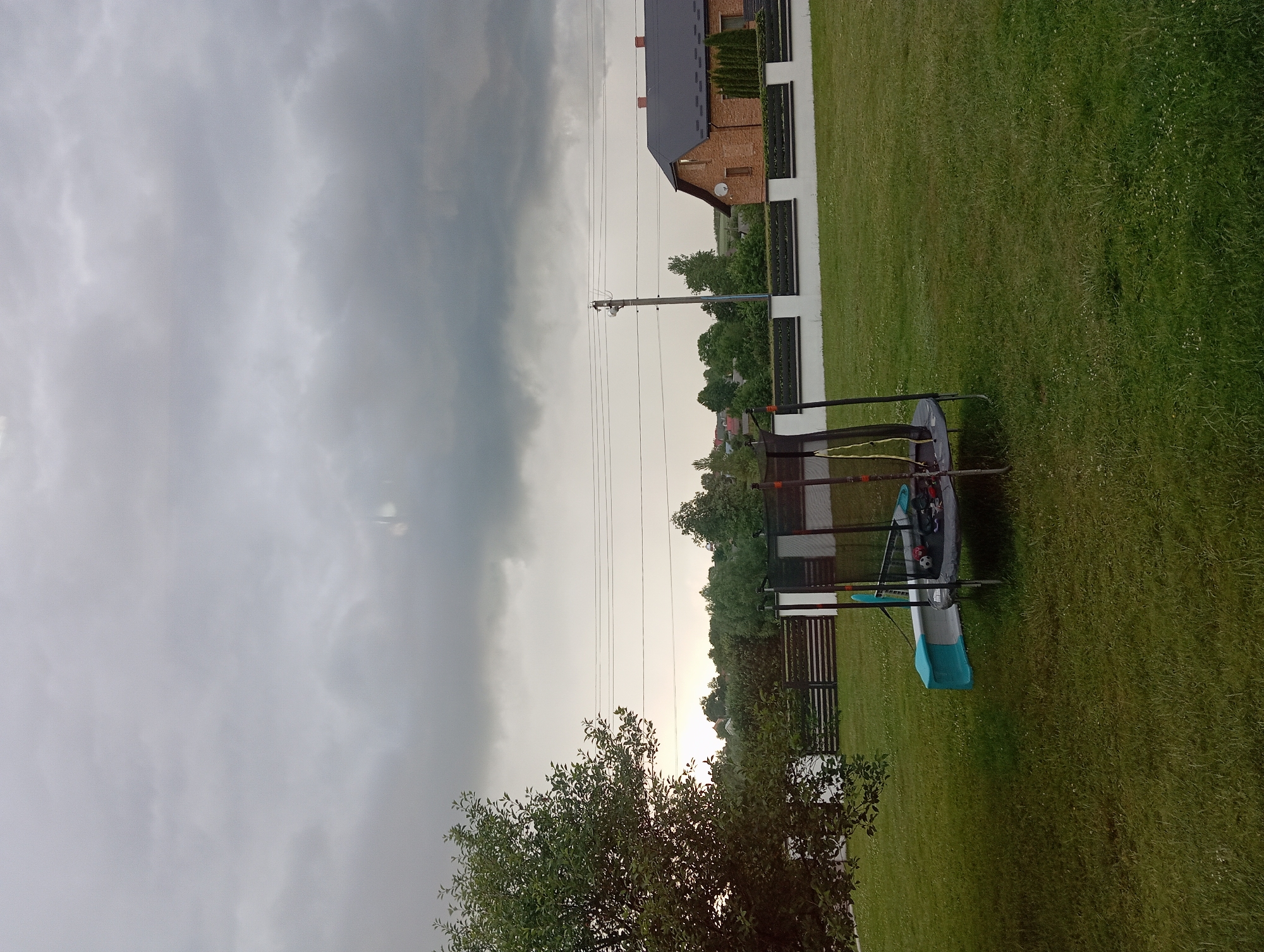 The image captures a backyard scene in Hlynytsi, Ukraine, under a heavy, overcast sky. In the foreground, a trampoline with a safety net is visible, accompanied by a blue and white slide. The lawn is green and well-maintained, with some scattered small white flowers. A mature tree with dense foliage stands to the left. In the mid-ground, a low white fence with horizontal dark elements separates the yard from a slightly elevated area with more trees and vegetation. Beyond this, a brick house with a dark roof and red chimney pots is situated on the right, with power lines and a utility pole visible in the distance. The overall impression is of a rural or suburban residential area on a cloudy day.