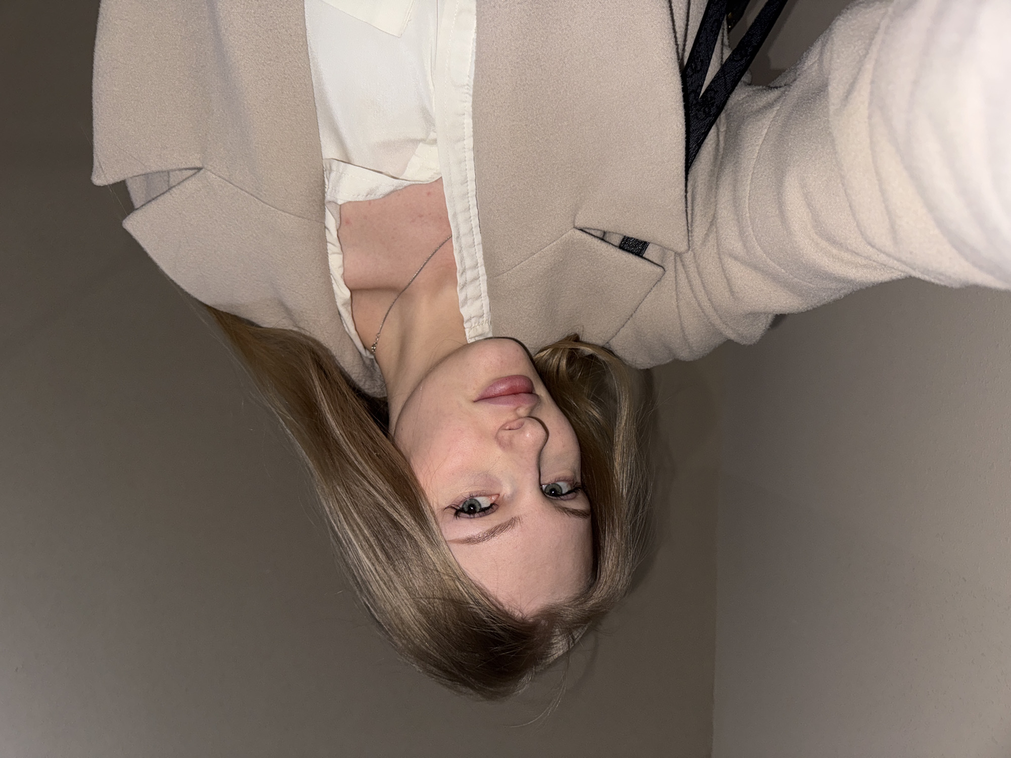 A young woman with light brown hair and light-colored eyes is depicted in a close-up, waist-up shot. Her head is tilted slightly, and she looks directly at the camera with a neutral to slightly serious expression. She is wearing a cream or light beige blazer over a white collared shirt, with a thin silver chain visible around her neck. Her hair is styled loosely, falling around her face and shoulders. The background is a plain, light grey wall, suggesting an indoor setting. The lighting appears dim, possibly indicating an evening or low-light environment. No specific location cues for Perm, Russia are visible, nor is any text present in the image. The composition suggests a selfie taken from a slightly elevated angle.