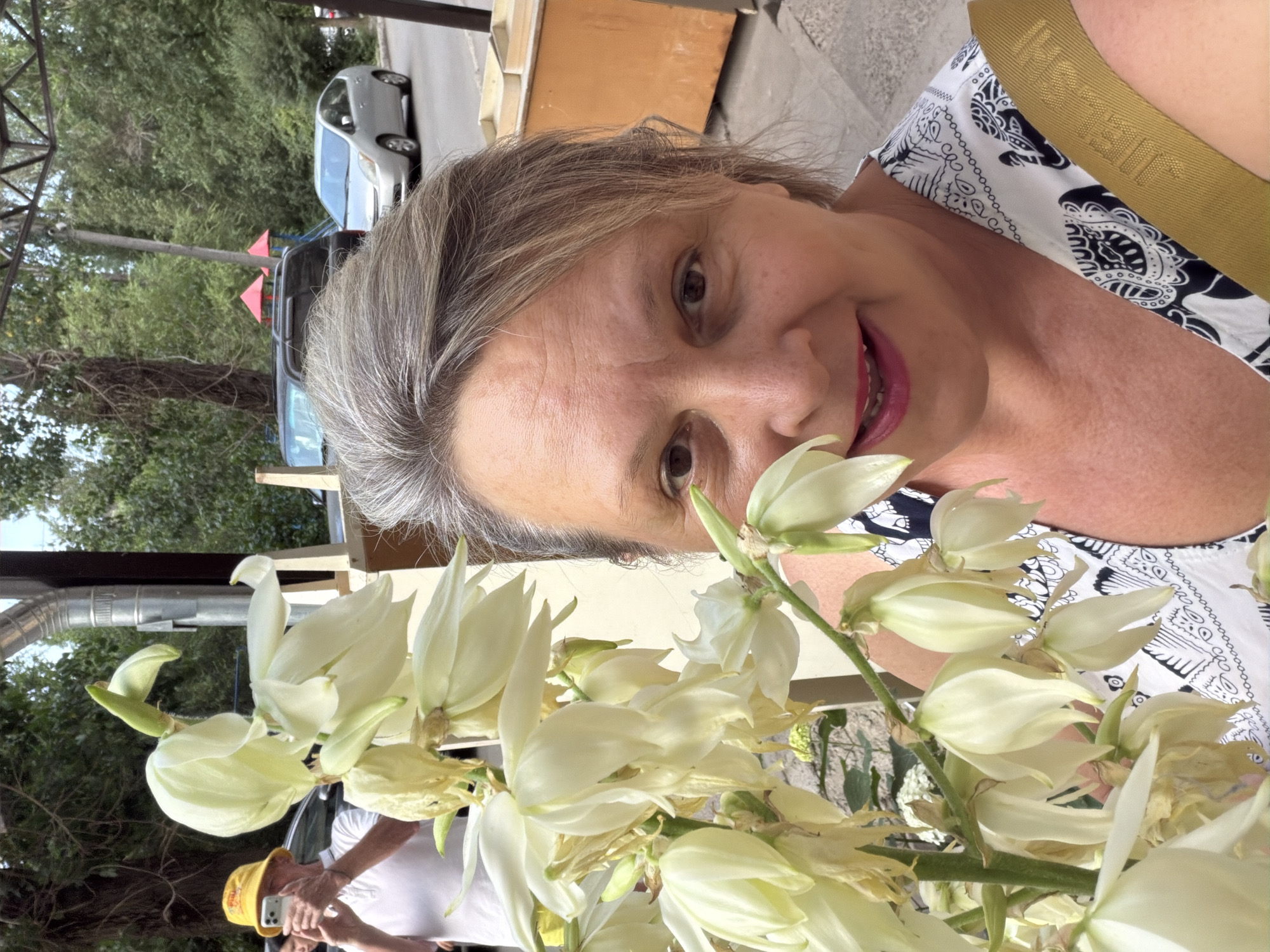A medium shot captures a Caucasian woman in her 50s with short gray hair and fair skin, taking a selfie outdoors. She has brown eyes and is wearing lipstick. She is partially obscured by a stalk of white flowers. The woman is wearing a black and white patterned top with a gold strap across her shoulder. In the background, a person in a yellow hat is visible, holding a smartphone, seemingly taking a photo. A black SUV and a silver car are parked on a street. The scene appears to be on a street with trees and a building with an orange facade.