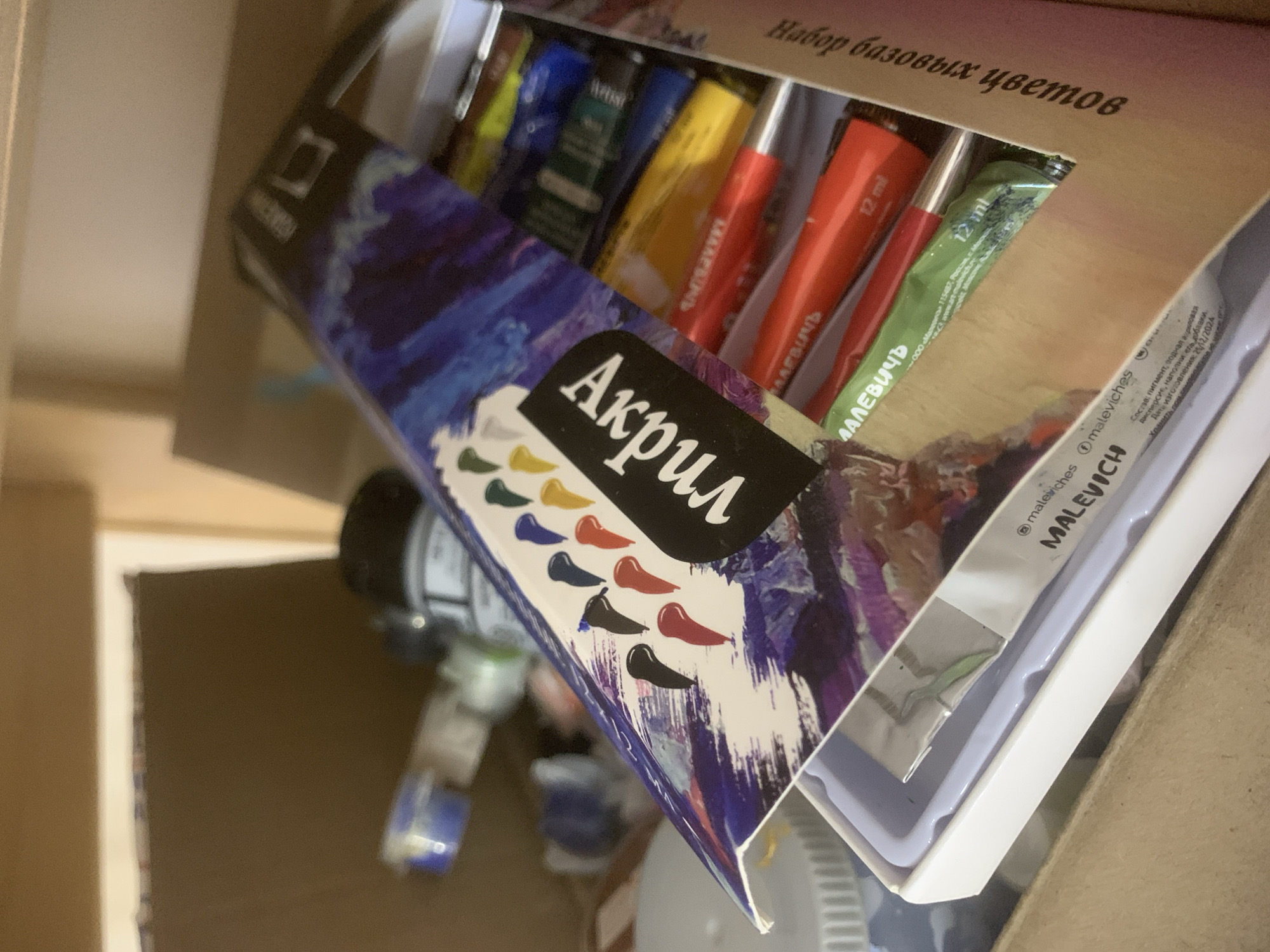 The image shows a box of "Malevich" brand acrylic paints ("Акрил") in a cardboard box. The paint tubes are visible inside, with Russian text on the box and tubes including "Набор базовых цветов" ("Basic set of colors"). The packaging includes the manufacturer's name, "MALEVICH," and other information in Russian, including the date of manufacture, 25/12/2024, and an address. The tube is 12 ml. Other items are also visible inside the cardboard box. The scene is indoors. Izhevsk is not mentioned in the image.