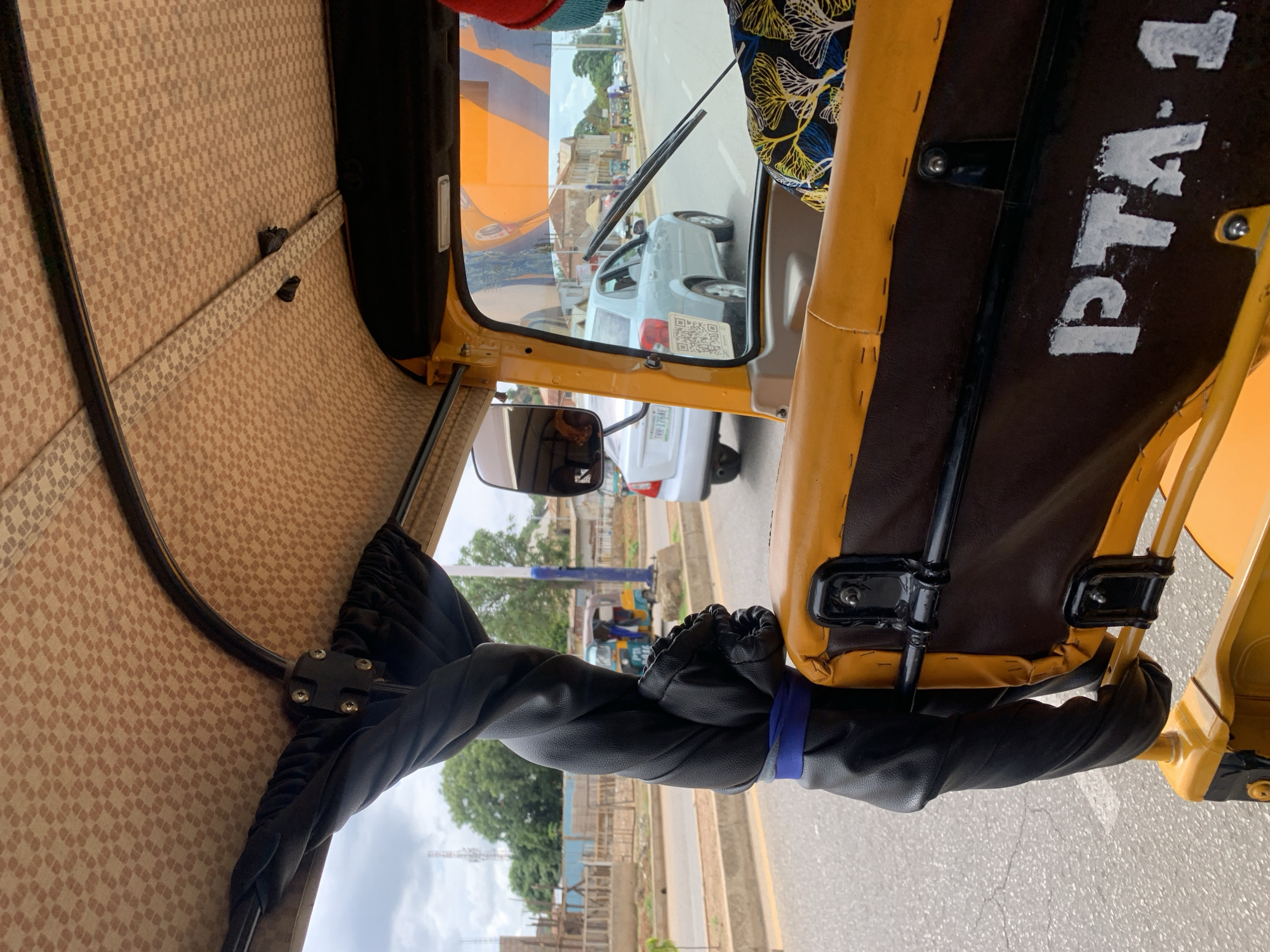 The image depicts an urban street scene in Kabong, Nigeria, viewed from the interior of a yellow and brown three-wheeled vehicle (likely a 'keke napep'). The perspective is from the front passenger side, looking forward and slightly right.
The vehicle's interior features a patterned light brown fabric roof lining with small, repeating dark brown square motifs. A thick, black, twisted padded support column, secured with a blue band at its base, is prominent on the left. The internal frame structure is yellow. Below the driver's seat, a brown panel with yellow piping displays the distressed white alphanumeric text "PTA-1". The back of the driver is visible on the right, clad in a shirt with a blue, yellow, and white foliage pattern, and a knitted hat with red and light blue sections. A single windshield wiper arm rests vertically on the windshield, and a side-view mirror reflects a blurred interior element.
Through the windshield, an asphalt road is visible, with a raised concrete median strip separating traffic lanes. A silver sedan, identified by a large black and white QR code sticker on its rear bumper and a partially visible license plate "TRR-176AE", travels ahead in the same direction. Further down the left side of the road, another yellow and blue three-wheeled vehicle is present. The roadside on the left features a construction site with wooden scaffolding and partially built concrete structures. Green trees are interspersed along the street, and a blue utility pole is visible. The sky is overcast.