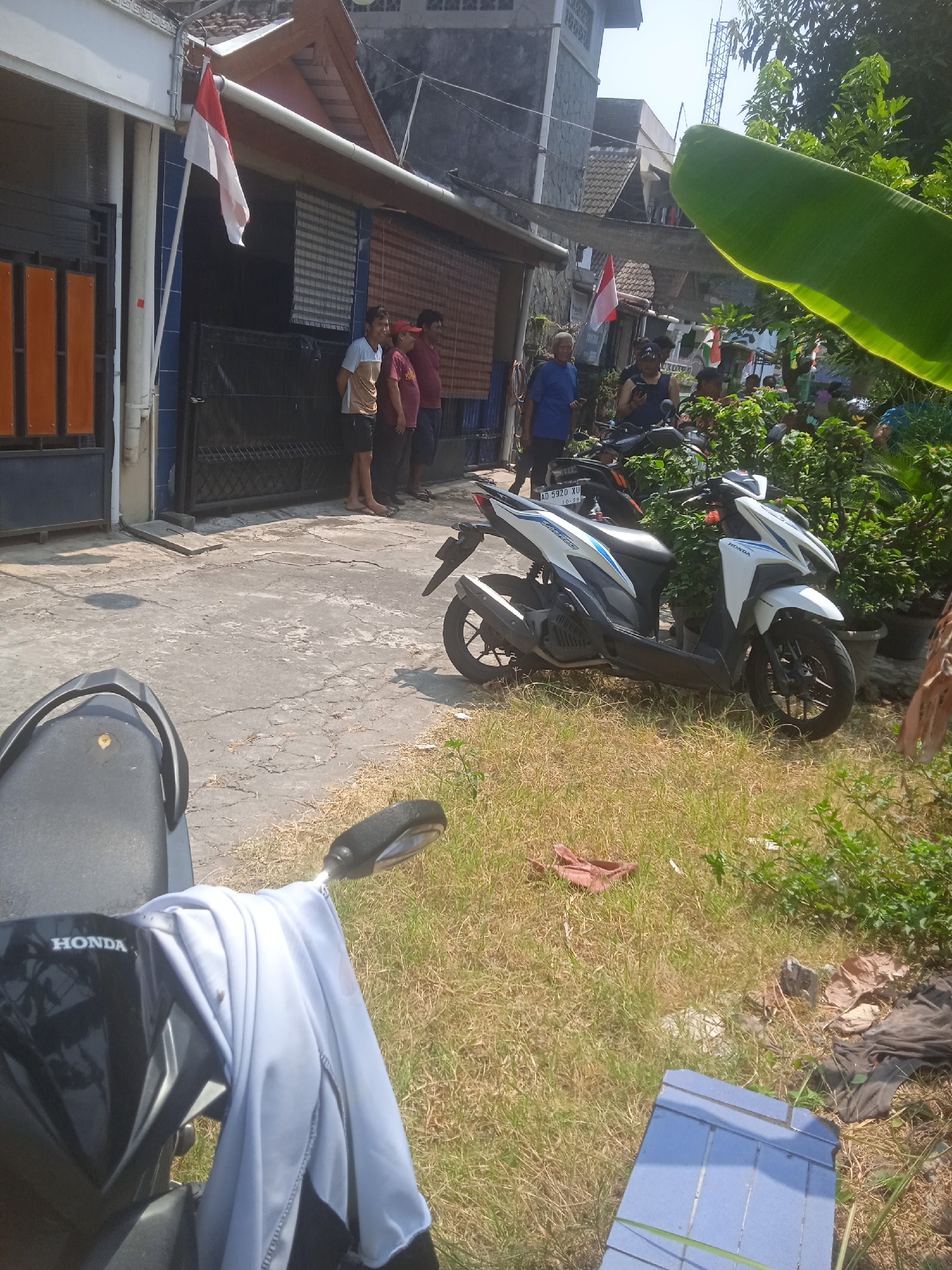 In Klodran, Indonesia, the image captures a sunlit street scene with several individuals standing near a building. A white and red Indonesian flag is prominently displayed, affixed to a white pipe extending from the building's facade. Three men are visible standing in a line, facing away from the camera. To their right, a white and blue scooter is parked, with a license plate that reads "AD 5920 XU". In the foreground, the back of a black scooter is visible, with a light blue garment draped over its handlebars. The ground is a mix of cracked concrete and dry grass. In the background, other buildings and dense foliage are present. The sky is clear and bright.