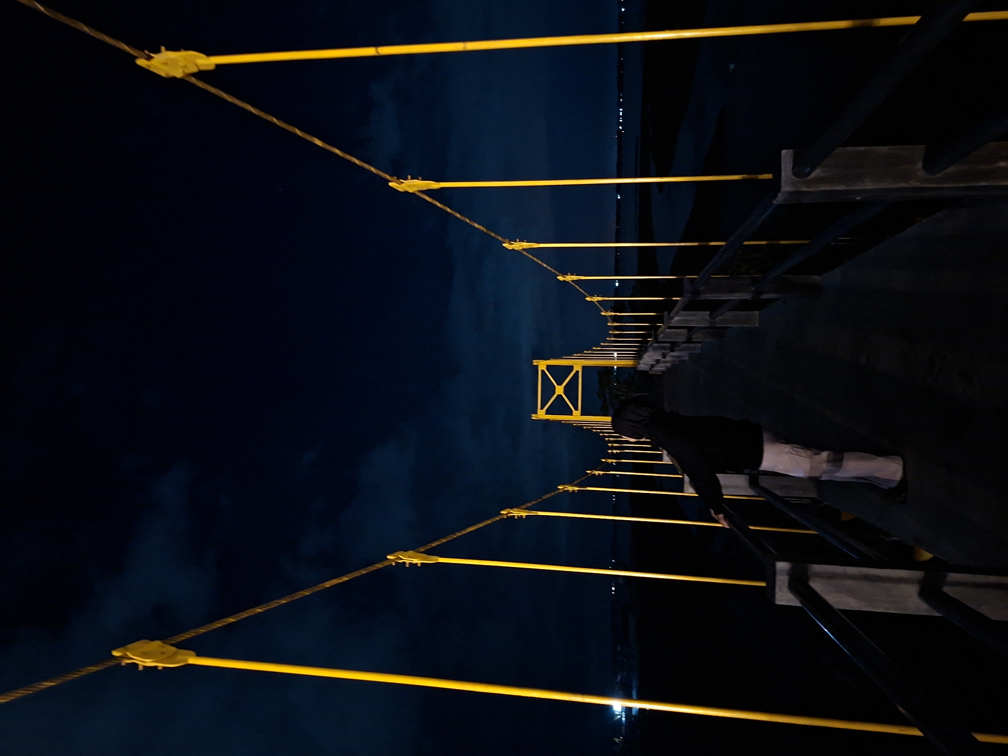 A low-angle, tilted night shot shows a pedestrian on the illuminated metal and cable suspension bridge, Crueng Geukueh, in Lhokseumawe, Indonesia. The person faces forward, holding the dark-colored metal railing on the right. The bridge’s yellow support cables and posts are visible, leading to a distant central tower. In the background is a dark horizon of land and water under a dark sky with scattered lights.
