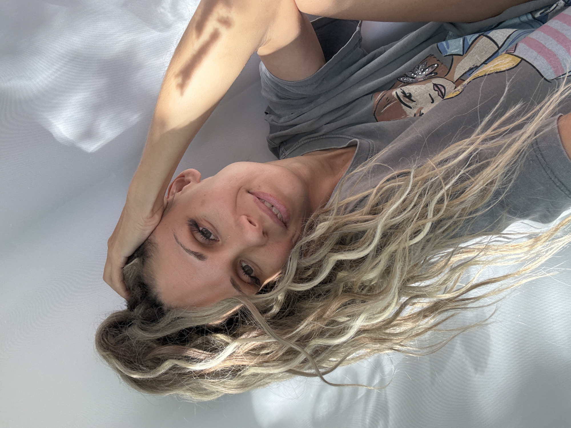 A woman with blonde, curly hair is captured in a selfie. She is wearing a grey t-shirt with a graphic print of a woman. Her right hand is resting on her head, and her left arm is bent with her hand resting near her face. The background is a white wall with light filtering through it, creating shadows. The image does not provide any information about the location in the city of Rusanovo, Russia.