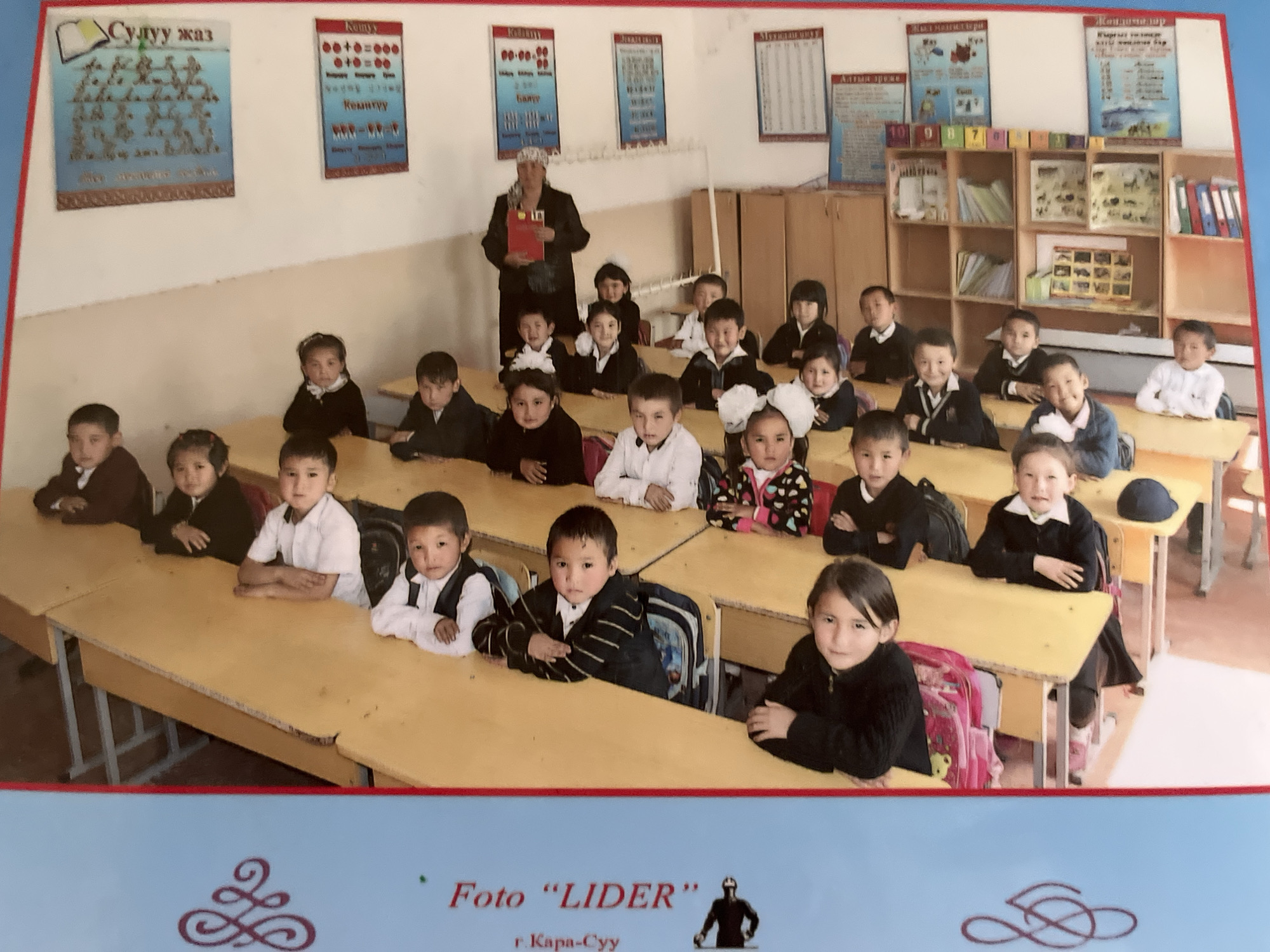 A group of young children, appearing to be primary school age, are seated at wooden desks in a classroom. They are mostly wearing dark-colored school uniforms with white shirts and some have ties. The children are positioned in rows, facing forward and looking at the camera. One child in the back is holding a red folder. The classroom has a light-colored wall with educational posters displayed, including one with what appears to be Cyrillic lettering and mathematical equations. There is also shelving with various items and binders. The image is taken from a slightly elevated angle, capturing the entire group and the classroom environment. A blue border with decorative motifs and text is visible on the left side of the image. The overall scene suggests a formal school photograph being taken.