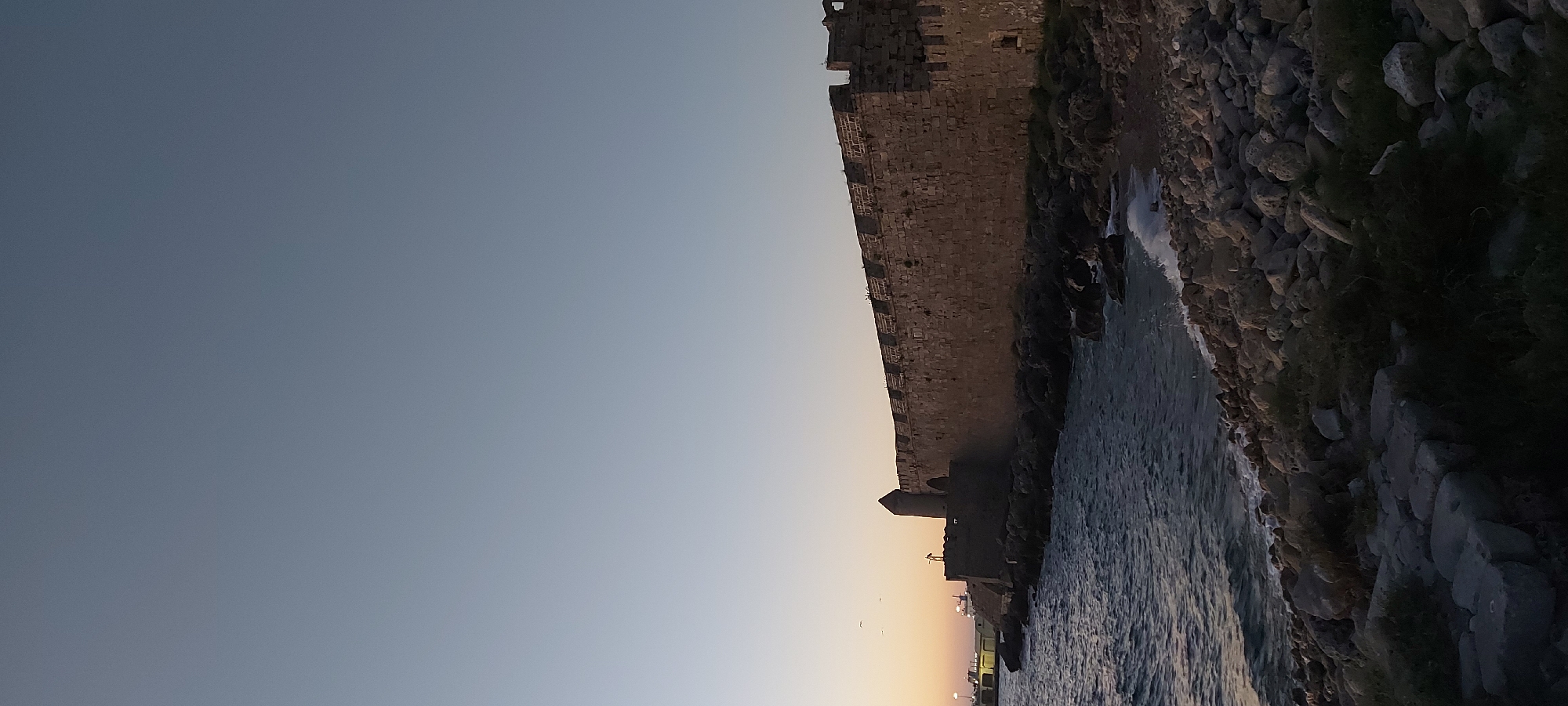 A long, ancient stone fortification wall runs horizontally across the frame, bordering a rocky shoreline. The wall, constructed from rough-hewn stones, has a crenellated top and features a small tower with a conical roof towards the left side. The sea, with gentle waves lapping against the rocks, fills the lower left portion of the image. The sky above is a clear, gradient of pale blue to a lighter hue near the horizon, suggesting either dawn or dusk. On the far left, a dock or pier structure is visible with some lights on. A few birds are in flight in the sky above the pier. The foreground is covered in a mix of large and small rocks, with some sparse vegetation growing among them.
