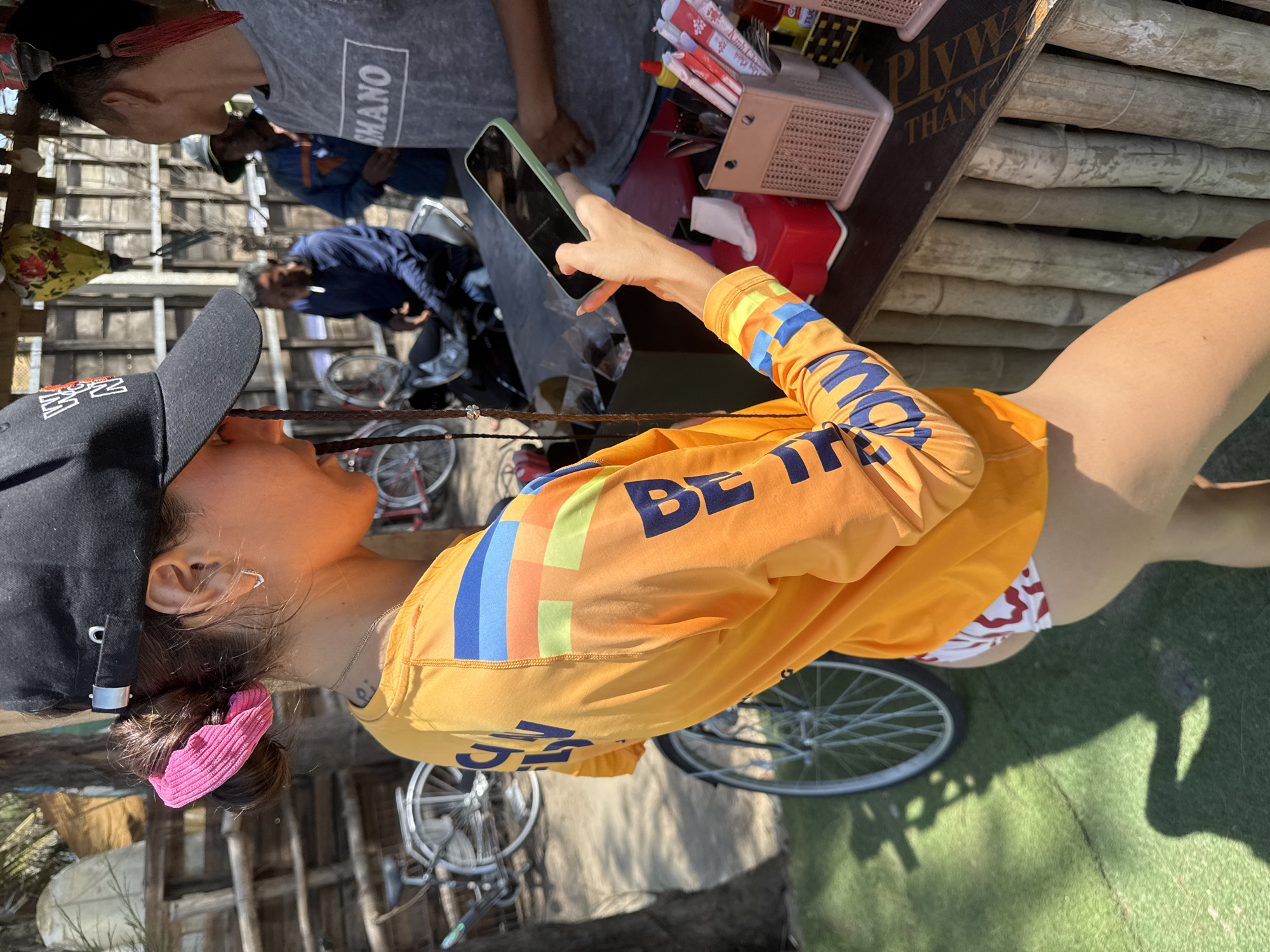 A woman with a black baseball cap and an orange long-sleeved shirt is using a smartphone. The shirt has "BE THE" printed in blue letters. She has a pink scrunchie in her hair. Several people and bicycles are in the background. The environment appears to be outdoors with dappled sunlight. The image was taken in Da Nang, Vietnam.