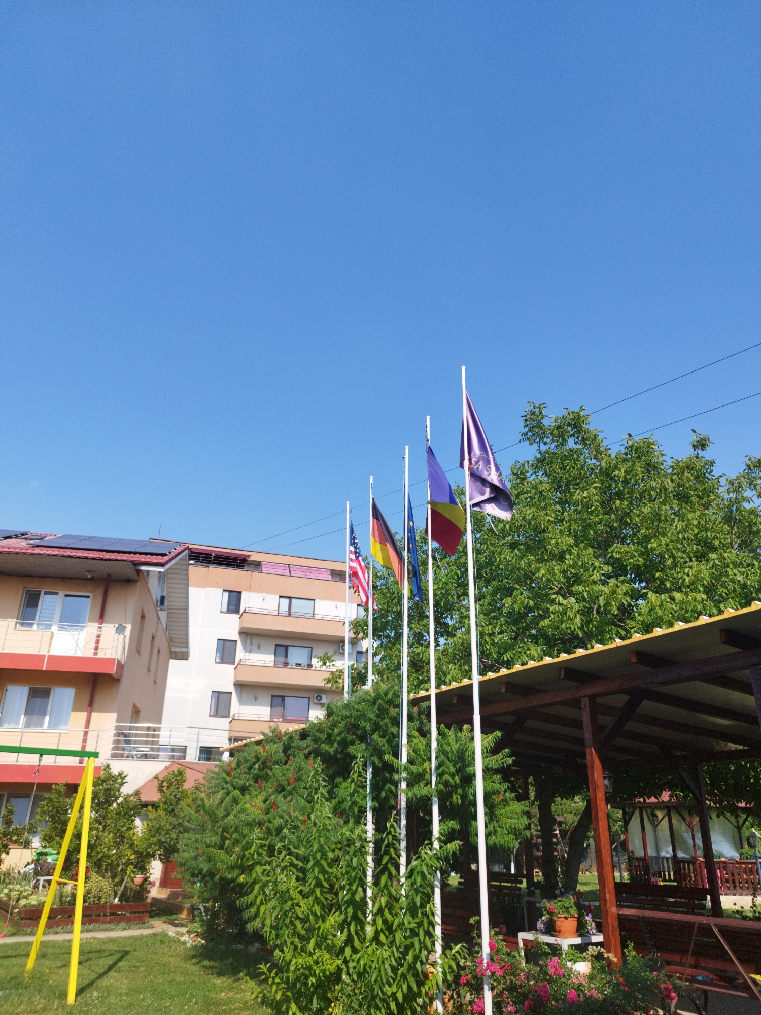 The image depicts a bright, sunny outdoor scene with a clear blue sky. In the foreground, a lush green lawn is visible, with a yellow swing set to the left. A line of flags flies on white poles: the flag of Germany, the flag of the United States of America, the flag of Romania, and a purple flag with white text that appears to read "HOUSE Sasha". Behind the flags, dense green trees form a backdrop. To the right of the flags, a wooden covered patio with a light-colored roof is present, featuring benches and potted plants. In the background, there are buildings, including a multi-story structure with balconies and solar panels on its roof, suggesting a residential or hospitality setting. The overall impression is one of a pleasant, possibly vacation or recreational, environment. The scene is likely located in Bucharest, Romania, as indicated by the presence of the Romanian flag and the architecture.