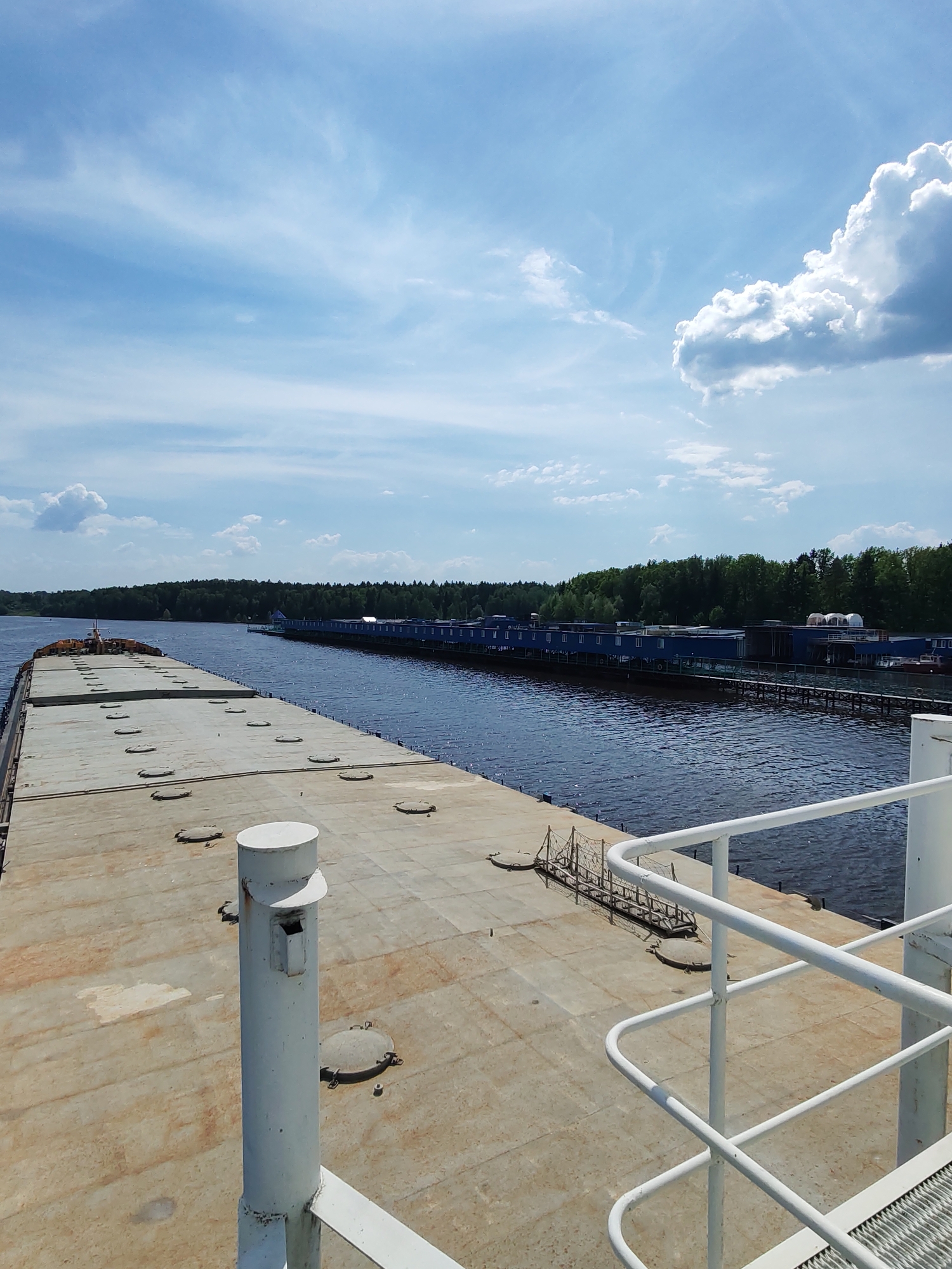 The image depicts a view from the deck of a barge or large vessel traversing a waterway. The deck is weathered and features circular access ports. A white railing and a cylindrical structure are visible in the foreground.
To the right, a shoreline with a long, blue structure with many windows is visible, likely a building or series of buildings on stilts along the water's edge. A dense forest lines the opposite bank.
The sky is blue with scattered white clouds. The water appears dark and calm. The location is Ermolino, Russia.