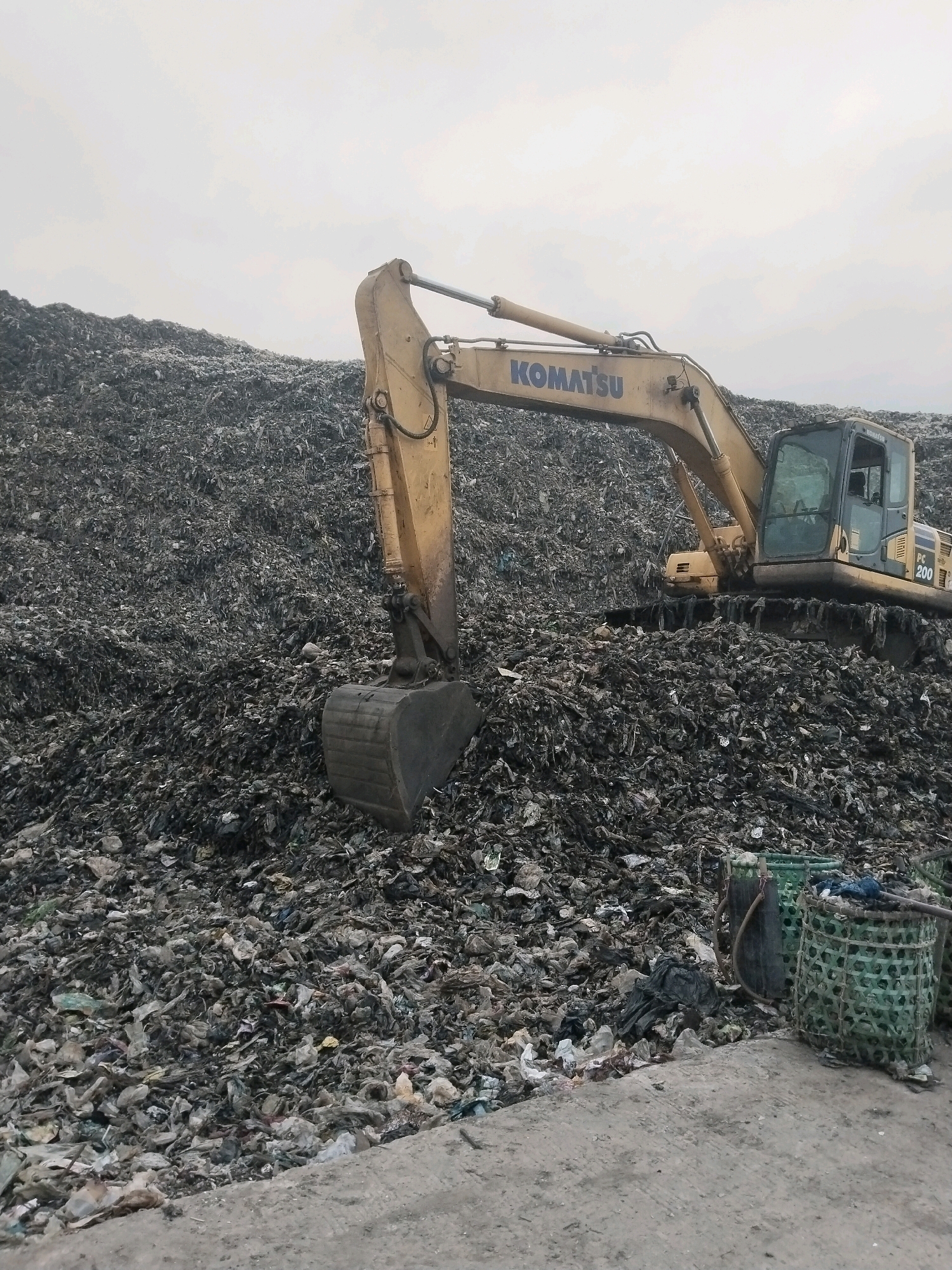 The image depicts a landfill site, likely in South Tangerang, Indonesia, characterized by a vast expanse of garbage. A yellow Komatsu excavator, identified by the "KOMATSU" logo and "PC 200" model number on its side, is positioned on the refuse pile, its bucket extended. The excavator appears to be actively working, though no specific action is clearly discernible. In the foreground, there is a concrete surface with several woven green baskets, some containing debris, and a dark cylindrical object, possibly a water drum, also filled with refuse. The sky is overcast, contributing to the dull and industrial atmosphere of the scene. The sheer volume of trash dominates the image, creating a landscape of waste.