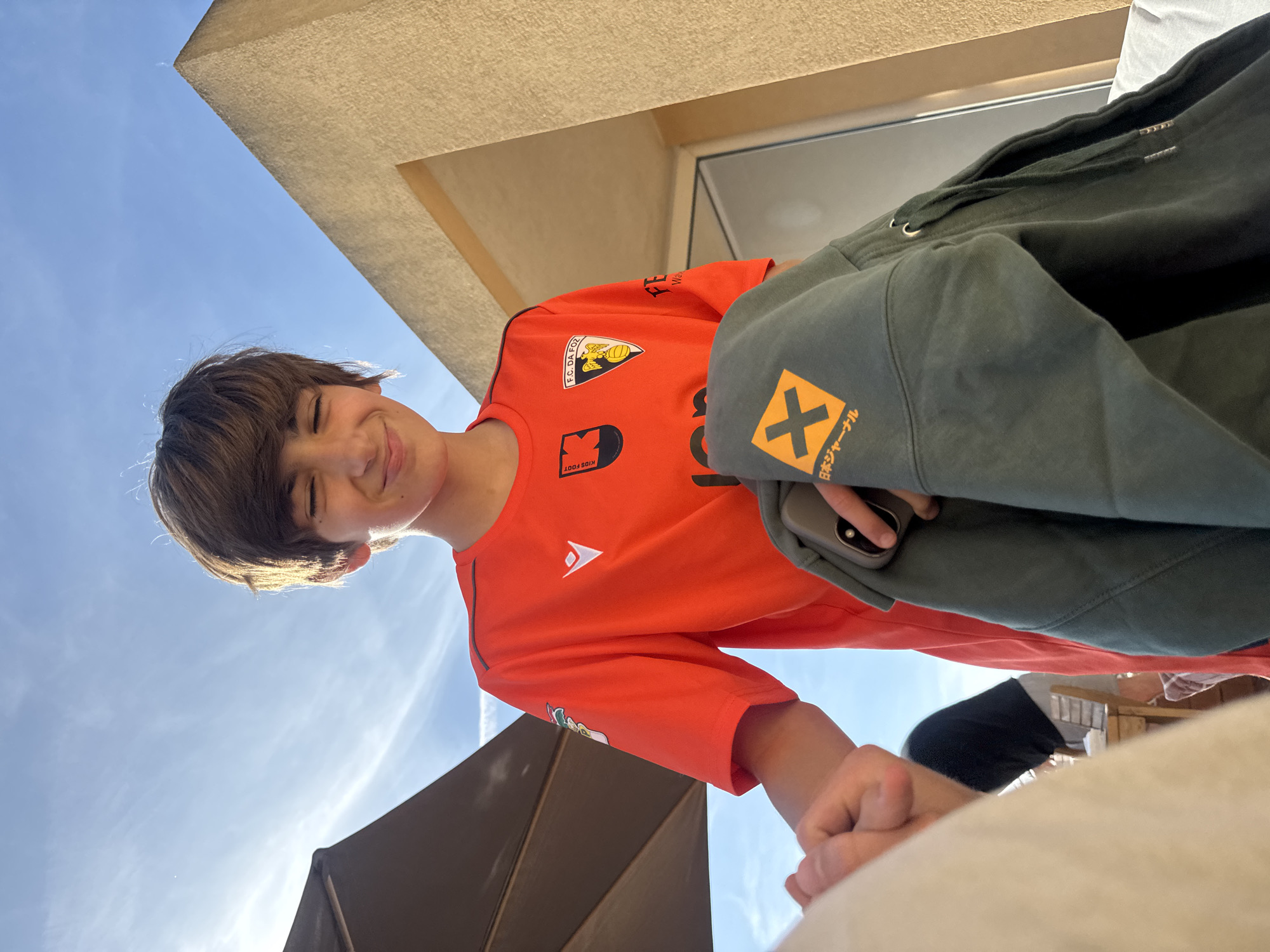 A young boy with short brown hair, wearing a bright orange t-shirt, is captured in an outdoor setting, looking down towards the camera with a joyful smile and squinting eyes, possibly due to direct sunlight. The image is taken from a low angle, looking upwards at the boy.
He is wearing a vibrant orange short-sleeved t-shirt featuring multiple logos. On his left chest, a crest displays an eagle-like bird holding a soccer ball with the text "F.C. DA FOR" above it. Further down the shirt, the text "KIDS FOOT" and other stylized graphics are visible. A dark green hoodie is draped over his right shoulder and arm, partially obscuring a smartphone he holds in his right hand. The hoodie's left sleeve prominently features a yellow 'X' symbol with the Japanese text "日本ジャーナル" (Nihon Journal) written beneath it.
The background consists of a clear, bright blue sky with some wispy clouds in the upper left. To the upper right, a light beige, modern building structure or overhang is visible, suggesting an urban or architectural environment. A dark, angular structure, possibly an umbrella or awning, occupies the lower right portion of the background, casting some shadow. The scene indicates a sunny daytime setting, likely in an outdoor area of Vila Nova de Gaia, Portugal, such as a patio or outdoor seating spot. The boy's expression conveys happiness and comfort.