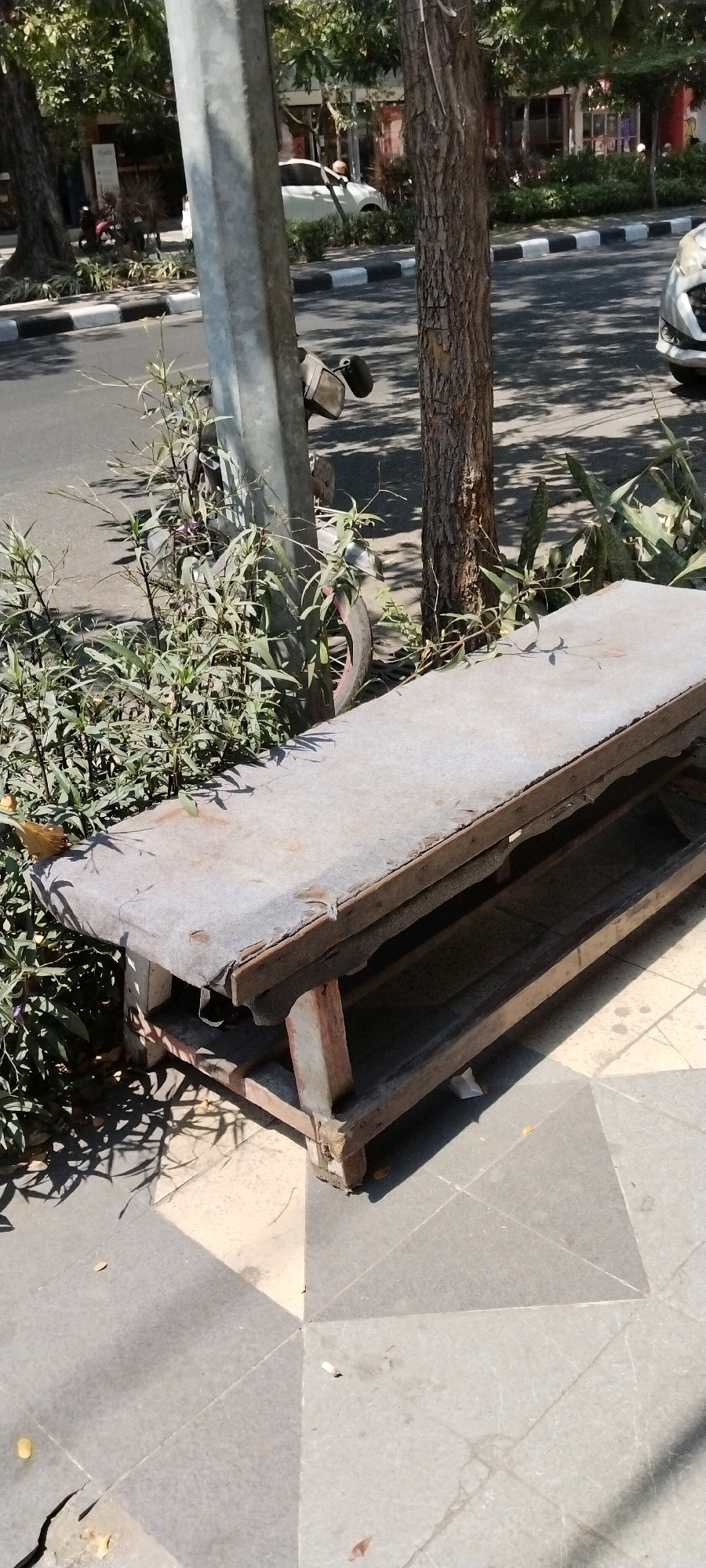 An aged, utilitarian wooden bench with a worn, grey fabric covering sits on a paved sidewalk next to a busy street. The bench is situated in front of lush green foliage and a large tree, with the dappled sunlight filtering through the leaves. In the background, a white car and a motorbike are visible, suggesting an urban environment. The pavement is made of grey tiles, some of which appear cracked. The scene is set on a bright, sunny day, casting distinct shadows across the ground. This photograph was taken on Jalan Taman Bungkul, Surabaya, Indonesia.