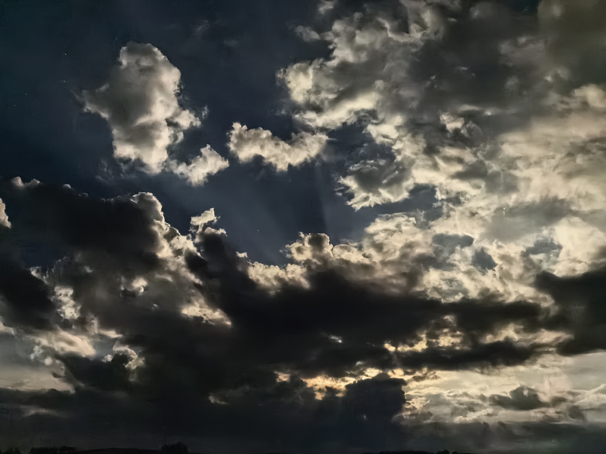 The image depicts a dramatic sky filled with cumulus clouds, illuminated by light that suggests either a setting or rising sun. The clouds range in color from dark grey to bright white, with varying degrees of opacity. Light shafts are visible, cutting through the cloud cover. The horizon is faintly visible at the bottom of the frame, indicating a landscape with minimal detail. The location is Unknown. There is a minimal amount of light illuminating what appears to be the ground in front of the clouds.